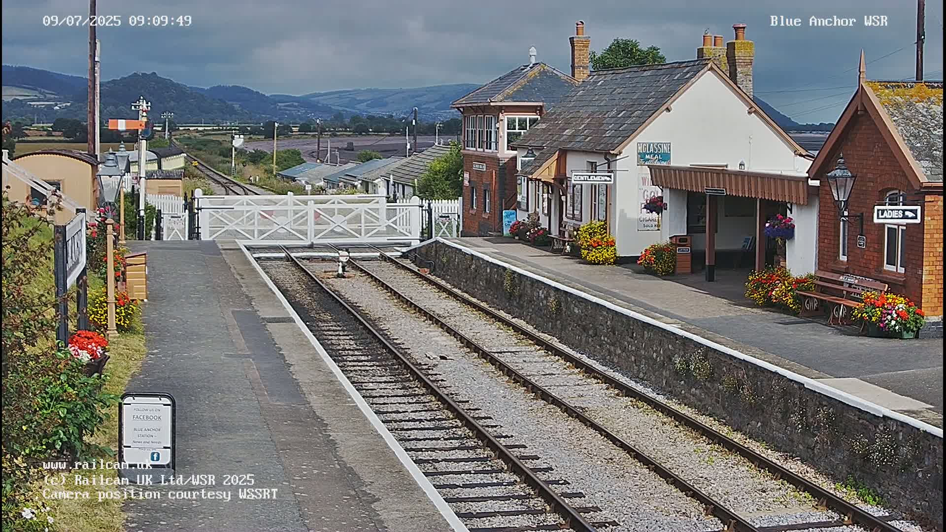 A train station with a signal gate, railway tracks, and several buildings, under a cloudy sky.