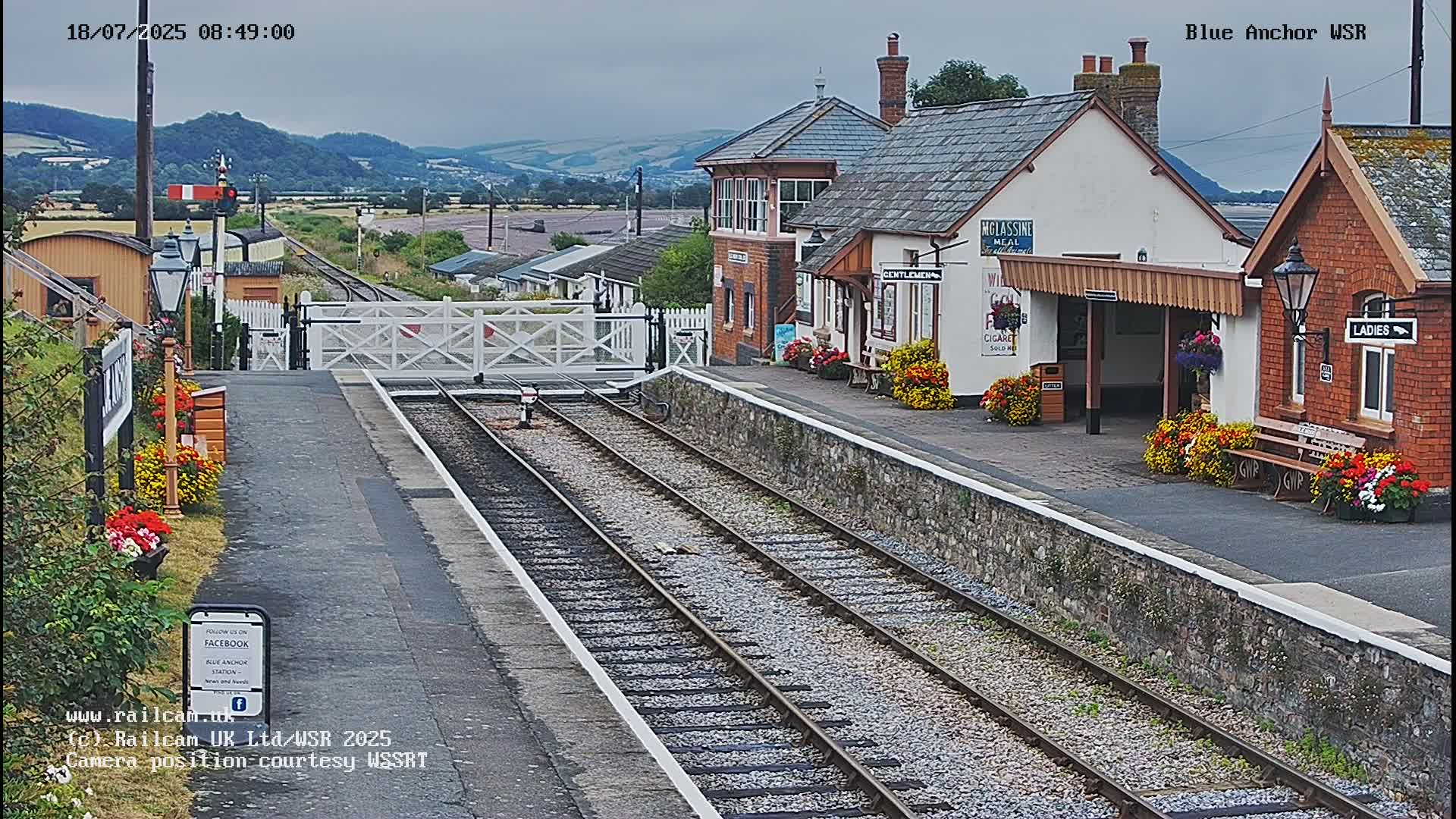 A railway station with a signal gate and train tracks, under an overcast sky, is surrounded by flower boxes and small buildings.