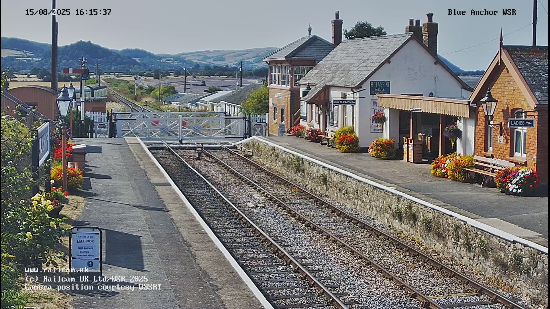 A railway station with a signal gate and train tracks, under an overcast sky, is surrounded by flower boxes and small buildings.