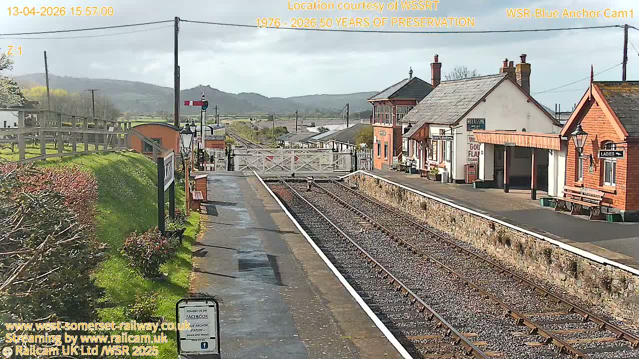 A railway station with a signal gate and train tracks, under an overcast sky, is surrounded by flower boxes and small buildings.