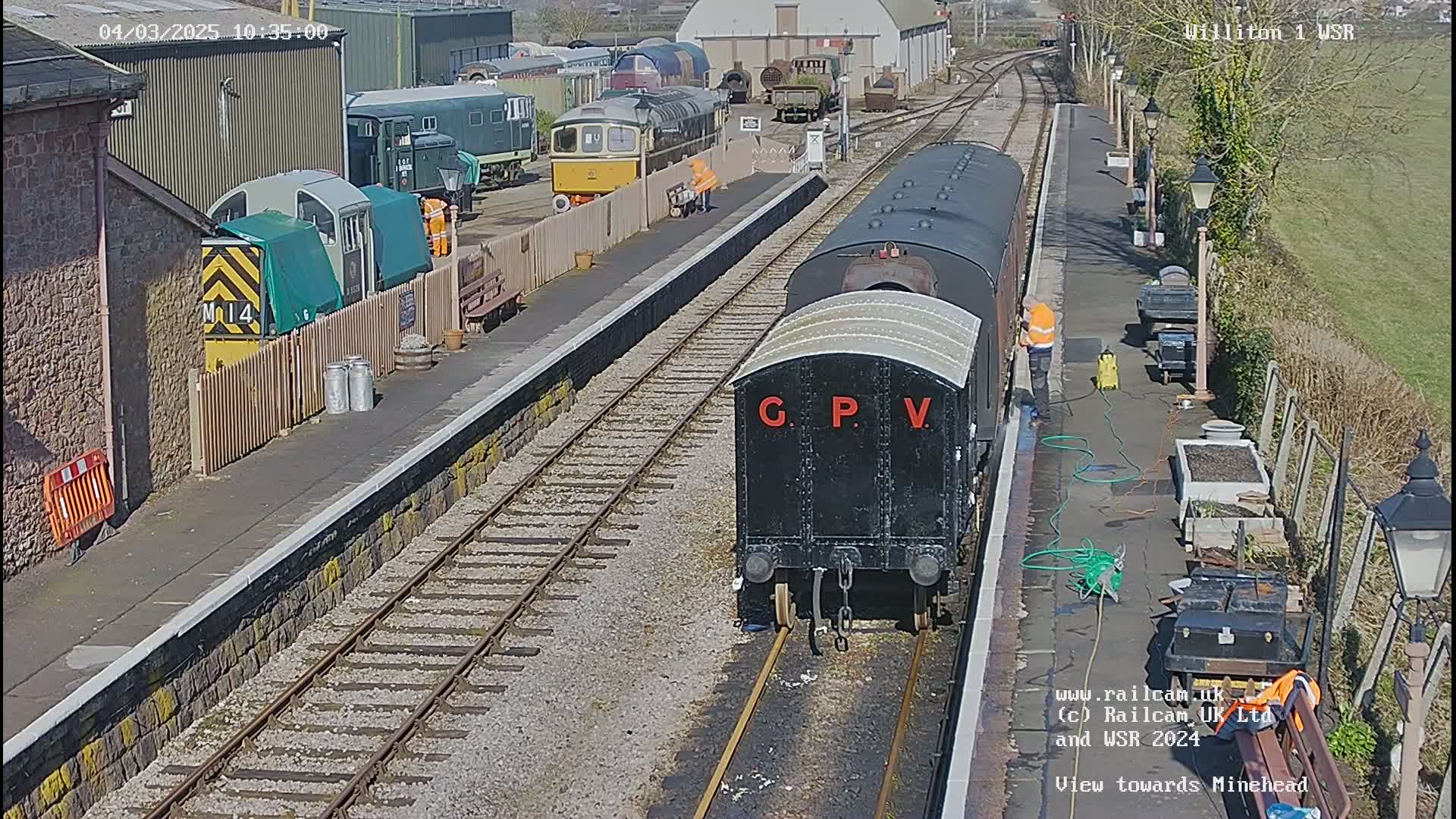 A black railway carriage labeled "G.P.V." is being cleaned at a train station on a sunny day, with other railway cars and buildings visible in the background.