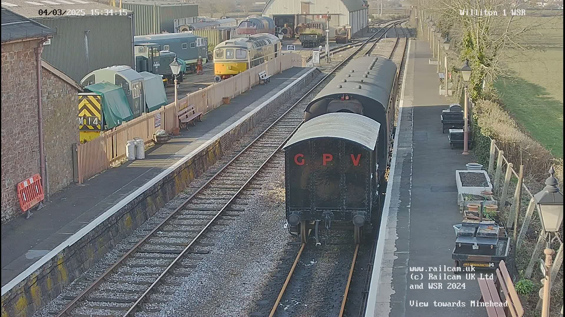 A black railway carriage with the letters GPV on its side sits on a railway track at a station on a sunny day, with other trains and railway buildings visible in the background.