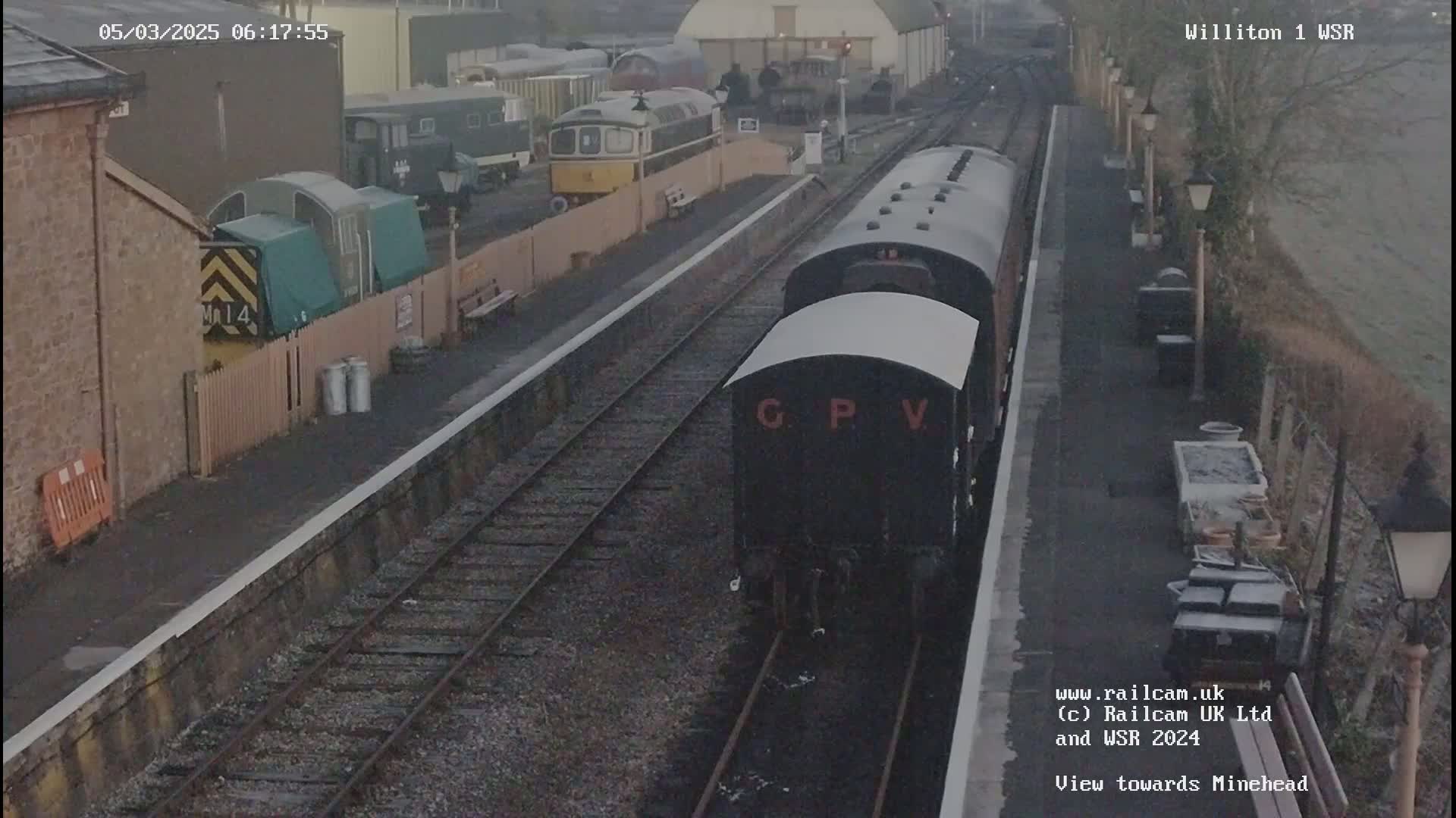 A train with a dark-colored engine and several cars is on a railway track at a station on an overcast day.