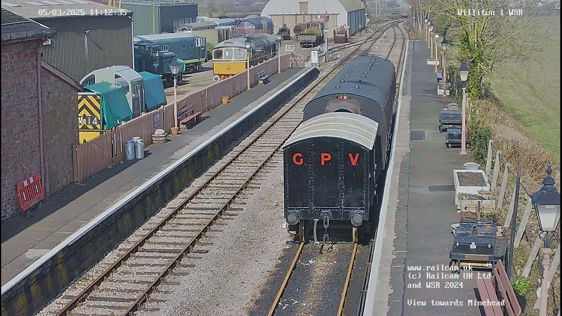 A black railway carriage labeled "G.P.V." sits on a sunny railway platform next to other railcars and railway buildings.