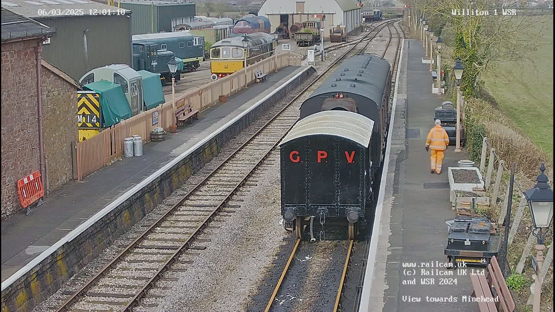 A black railway carriage sits on a track at a train station, where a person in an orange safety vest walks along the platform, with several other train cars parked nearby under a partly cloudy sky.