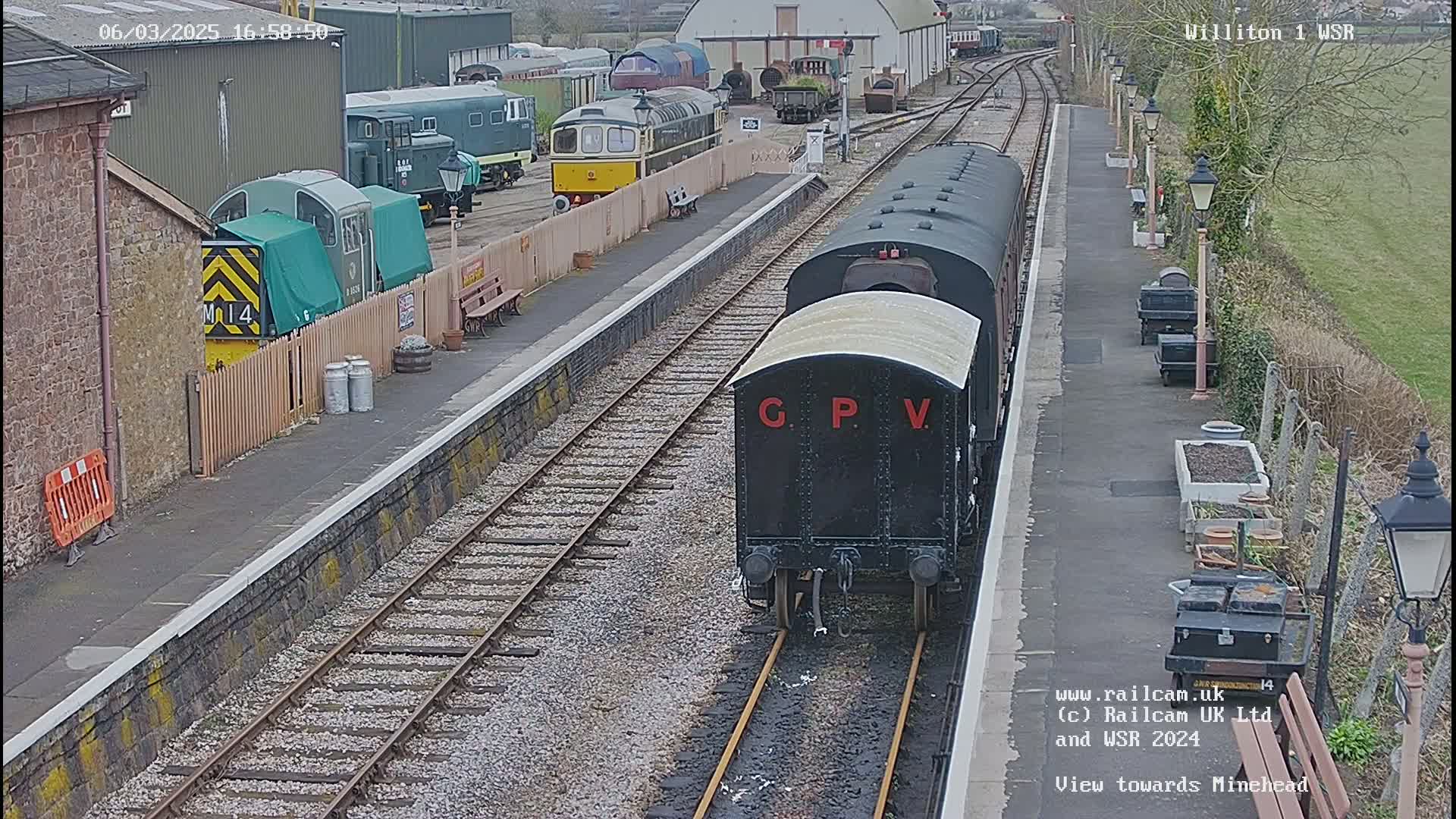 A black railway carriage labeled "GPV" sits on a track at a train station, with several other trains and buildings visible in the background on a partly cloudy day.