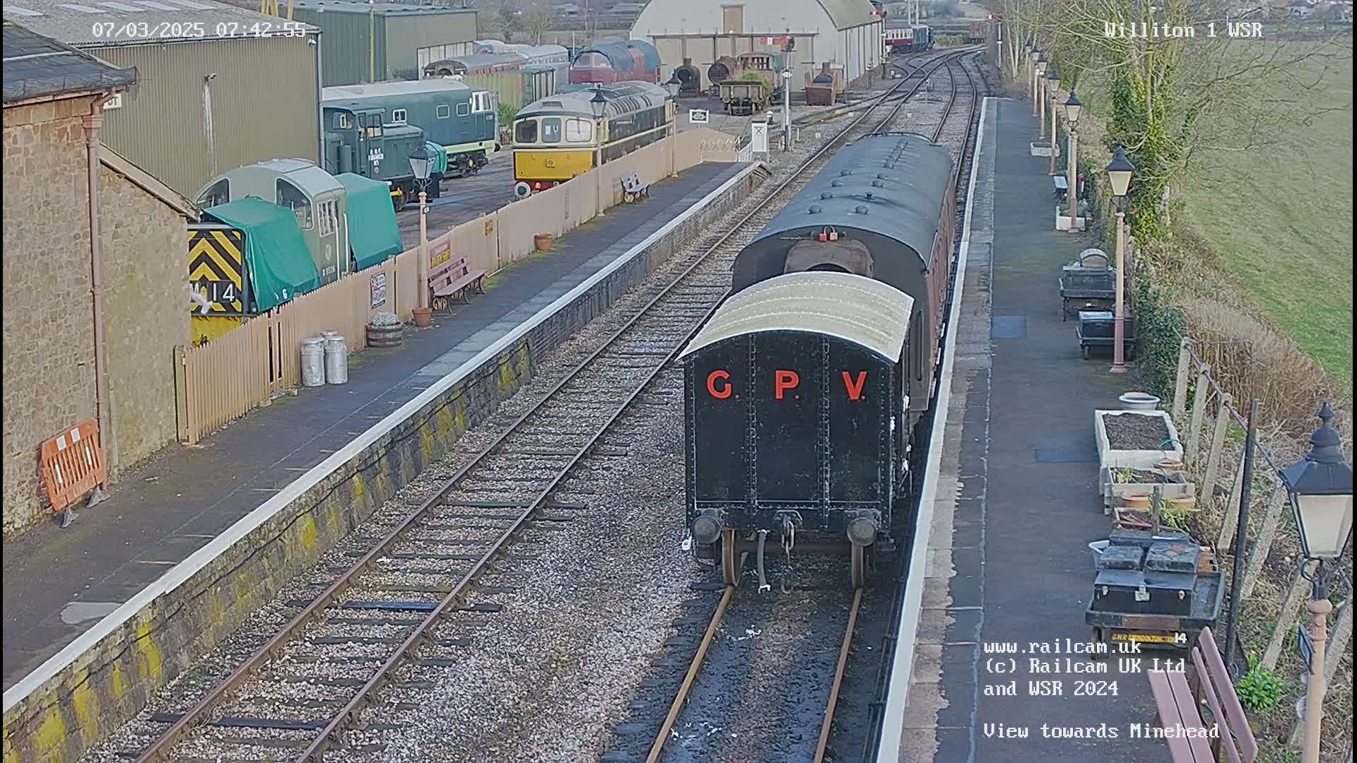 A black railway carriage labeled "G.P.V." is on the tracks of a train station with several other rail cars and buildings visible in the background, on a partly sunny day.