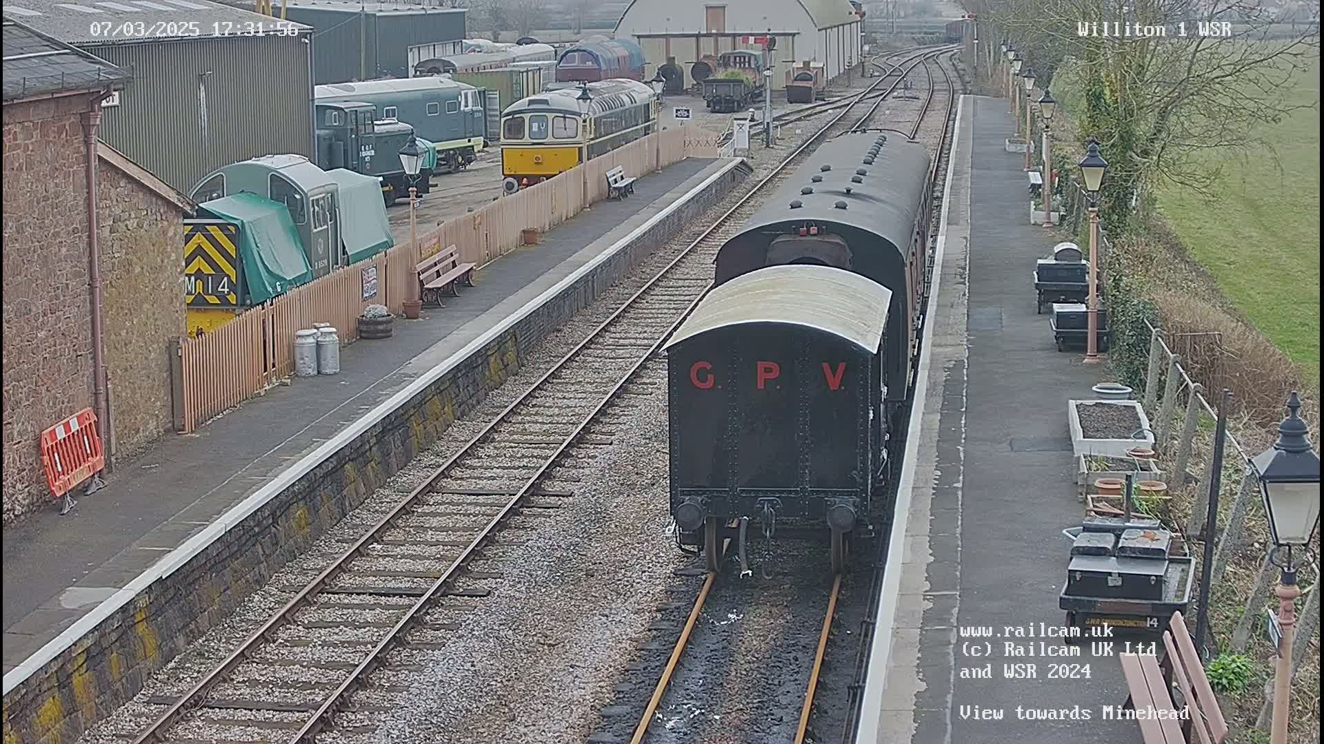 A dark-colored railway carriage approaches a station platform on a partly sunny day, with other train cars visible in the background.