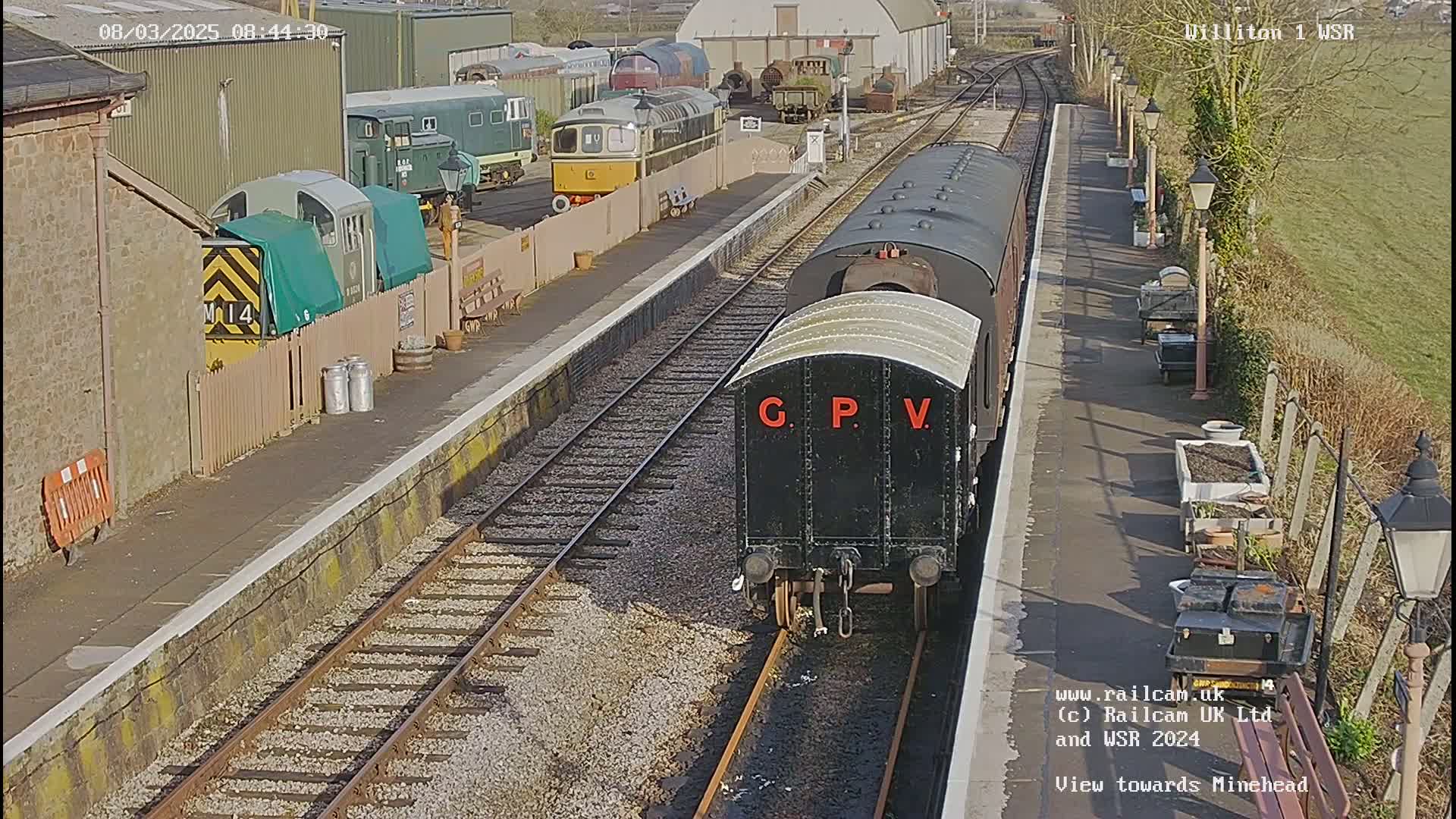 A black railway carriage labeled "G.P.V." is positioned on a train track next to a station platform on a sunny day, with various other trains and railway equipment visible in the background.