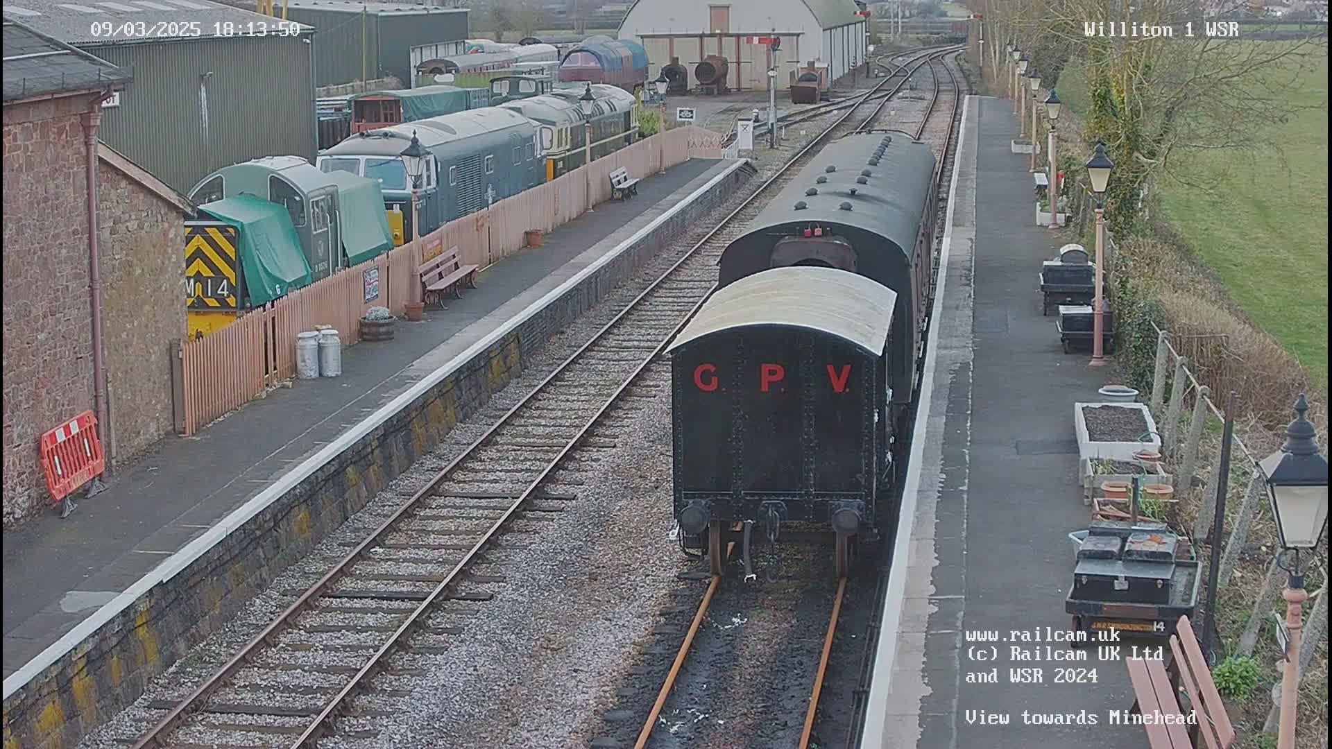 A black railway carriage labeled "G P V" is on a train track at a station, with several other train cars visible, in overcast daylight.