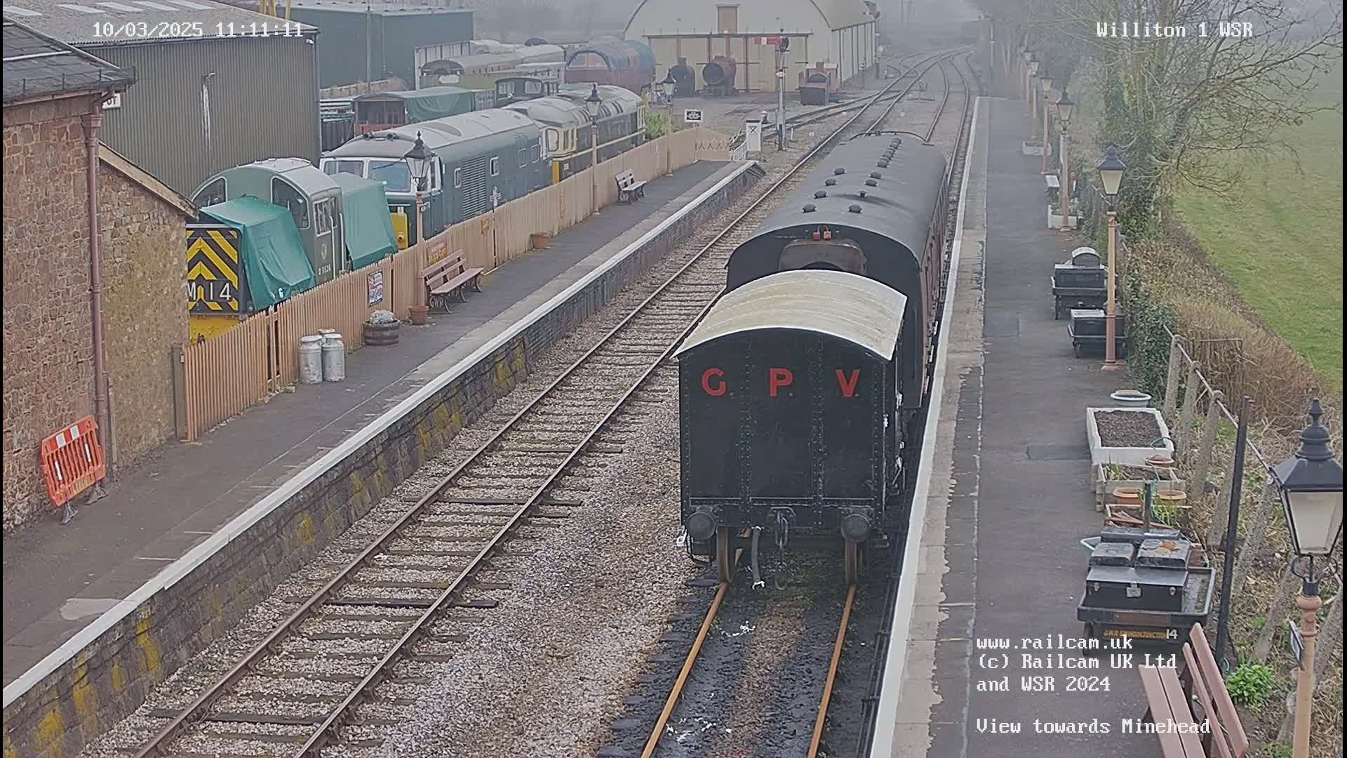 A black railway carriage labeled "GPV" is seen arriving at a train station on an overcast day, with other train cars and buildings visible in the background.