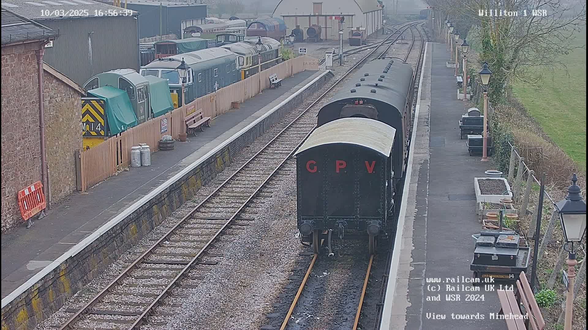 A black railway carriage labeled "GPV" approaches a train station on a cloudy day, with various other train cars visible in the background.