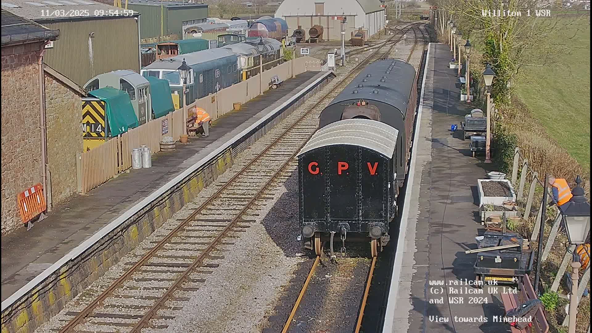 A black railway carriage labeled "G P V" sits on a railway track at a station on a sunny day, with other railway cars and buildings visible in the background.
