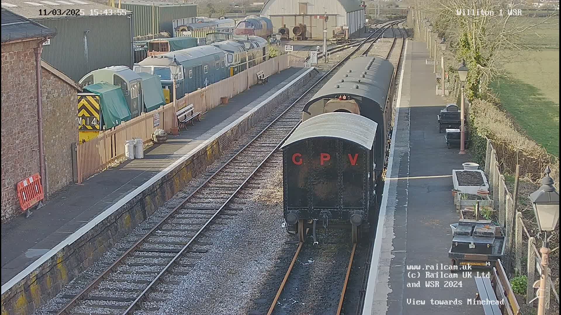 A black railway carriage is at a train station on a sunny day, with other train cars visible in the background.