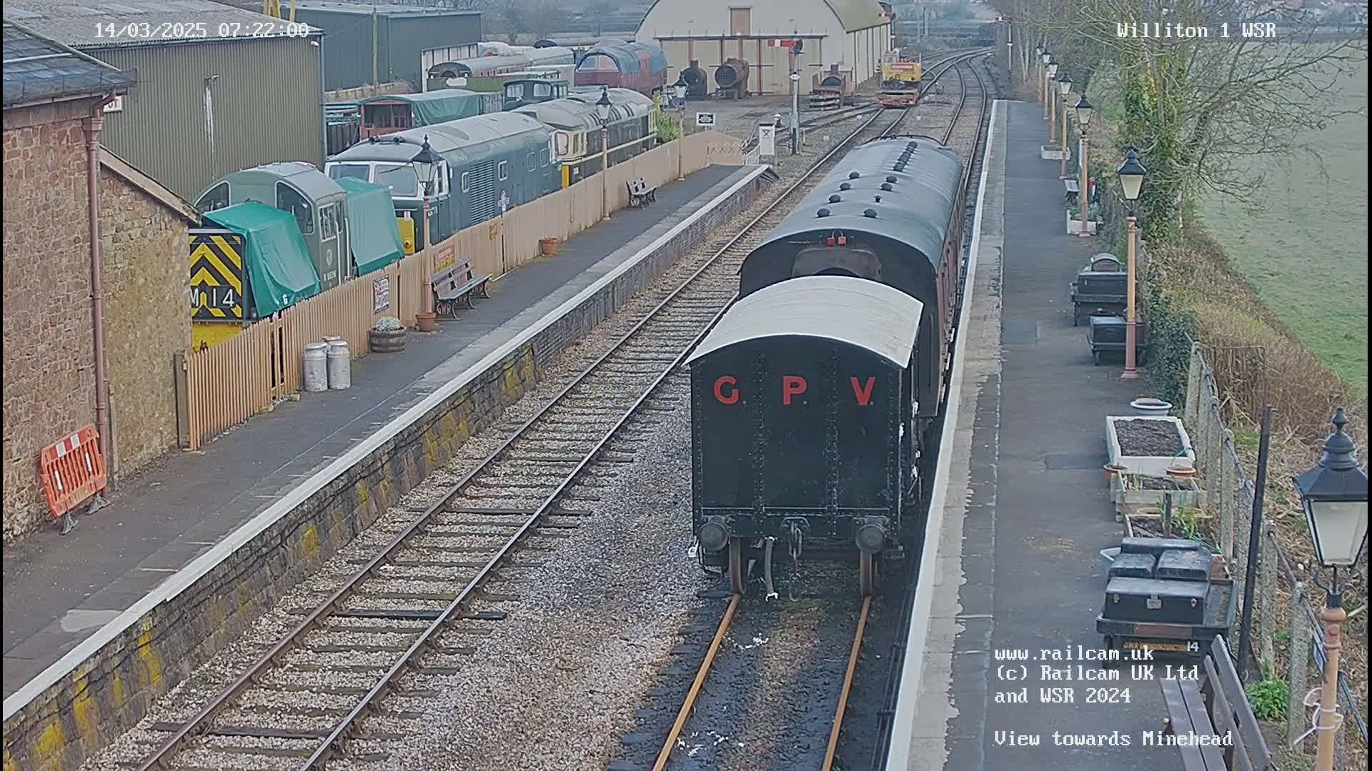 A black train car is on the tracks at a train station on an overcast day, with other train cars and a depot visible in the background.