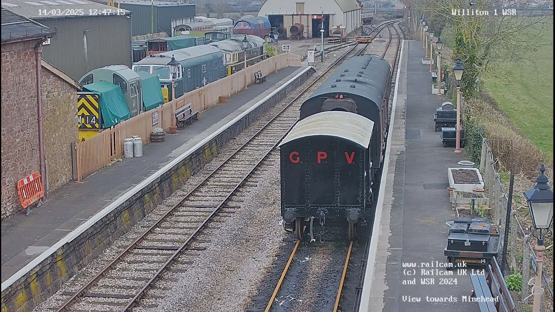 A dark-colored railway carriage labeled "GPV" is moving along tracks at a station, with several other train cars and a depot visible in the background on an overcast day.