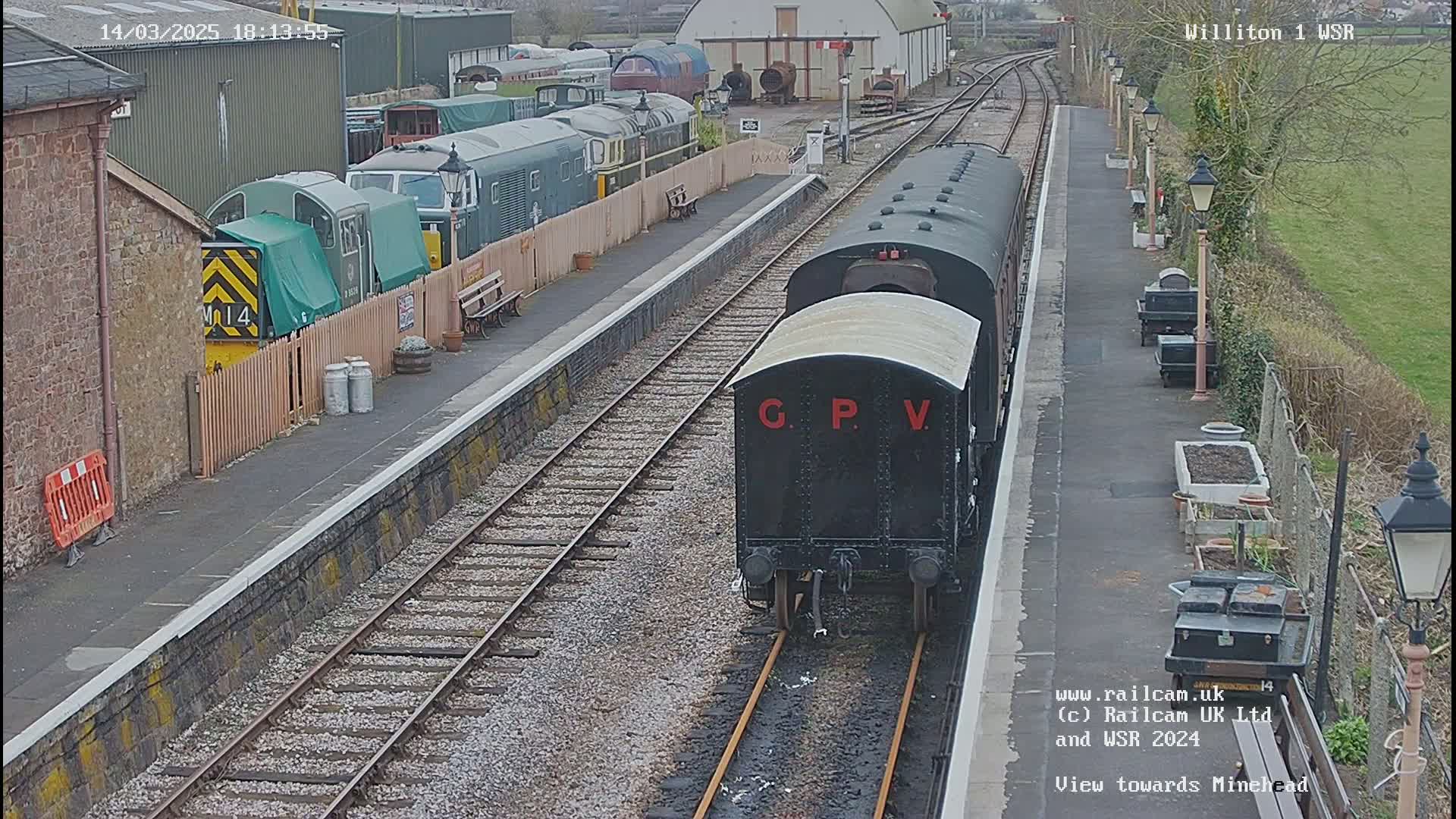 A black railway carriage labeled "G.P.V." is on a train track at a station on a partly cloudy day, with other train cars and buildings visible in the background.