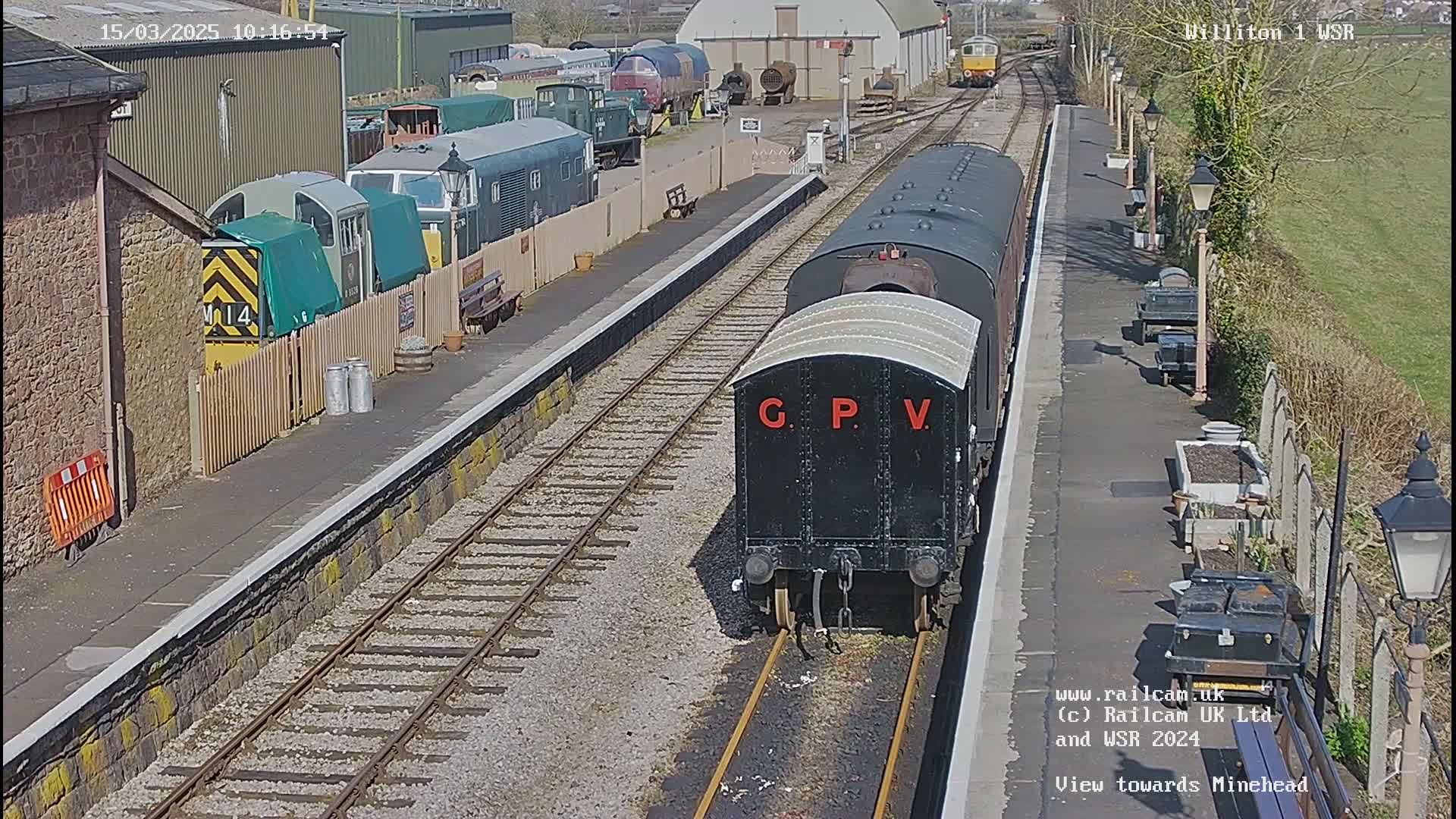 A black train car, labeled "G.P.V," sits on a railway track at a station on a sunny day.