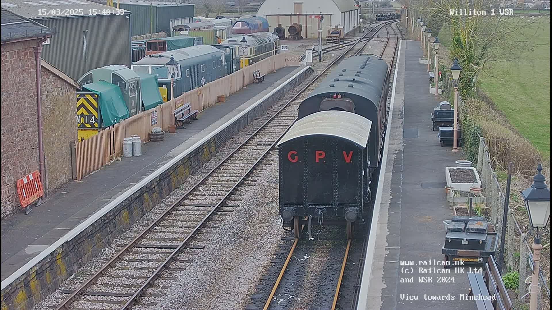 A black railway carriage labeled "G.P.V." is on a train track at a station with several other train cars and buildings in the background on a partly cloudy day.