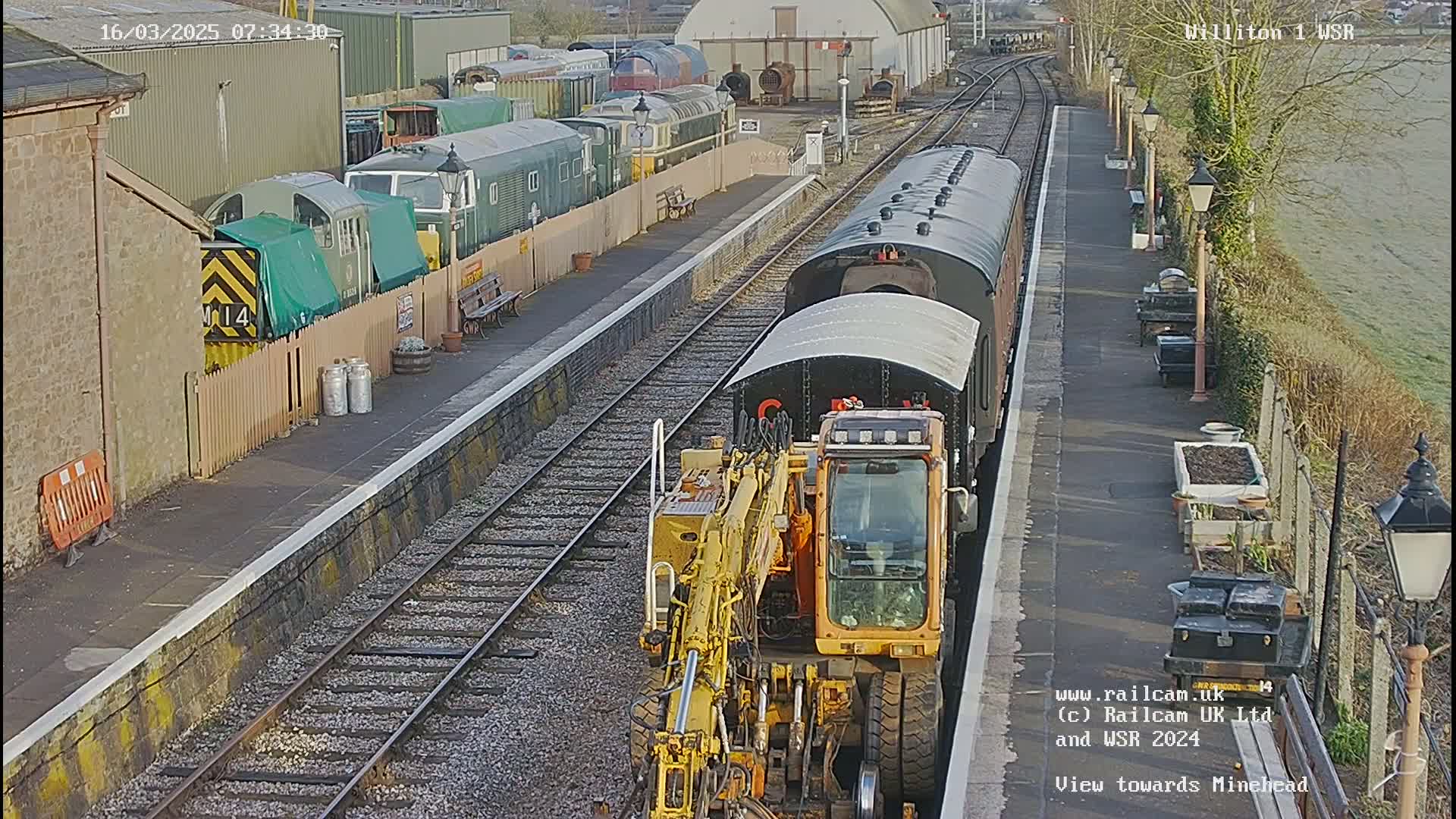 A yellow railway maintenance vehicle is positioned next to a dark-colored passenger train at a train station on a sunny day.