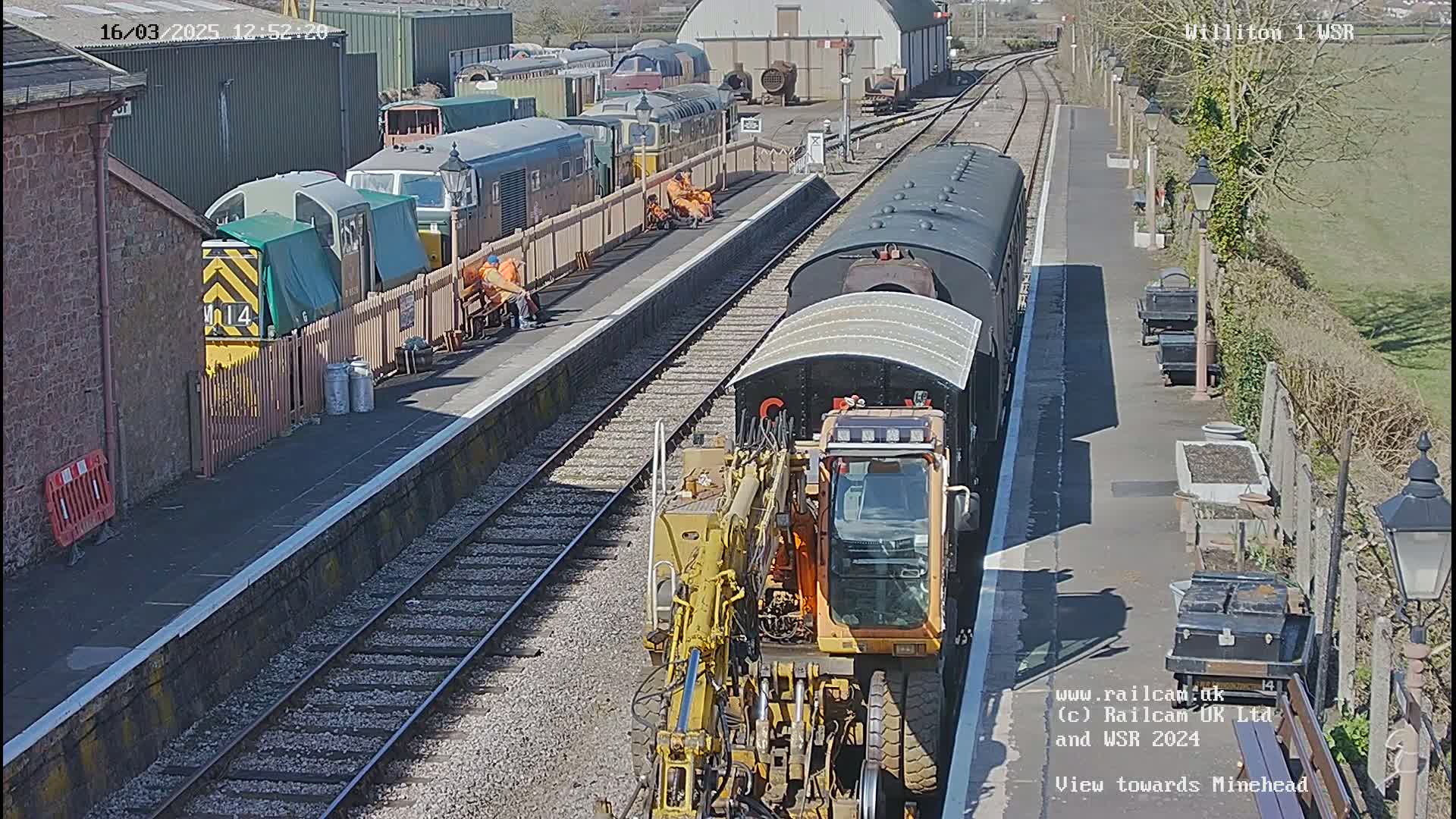 A yellow piece of railway maintenance equipment sits beside a dark-colored train on a sunny day at a train station with several other trains and railway cars visible in the background.