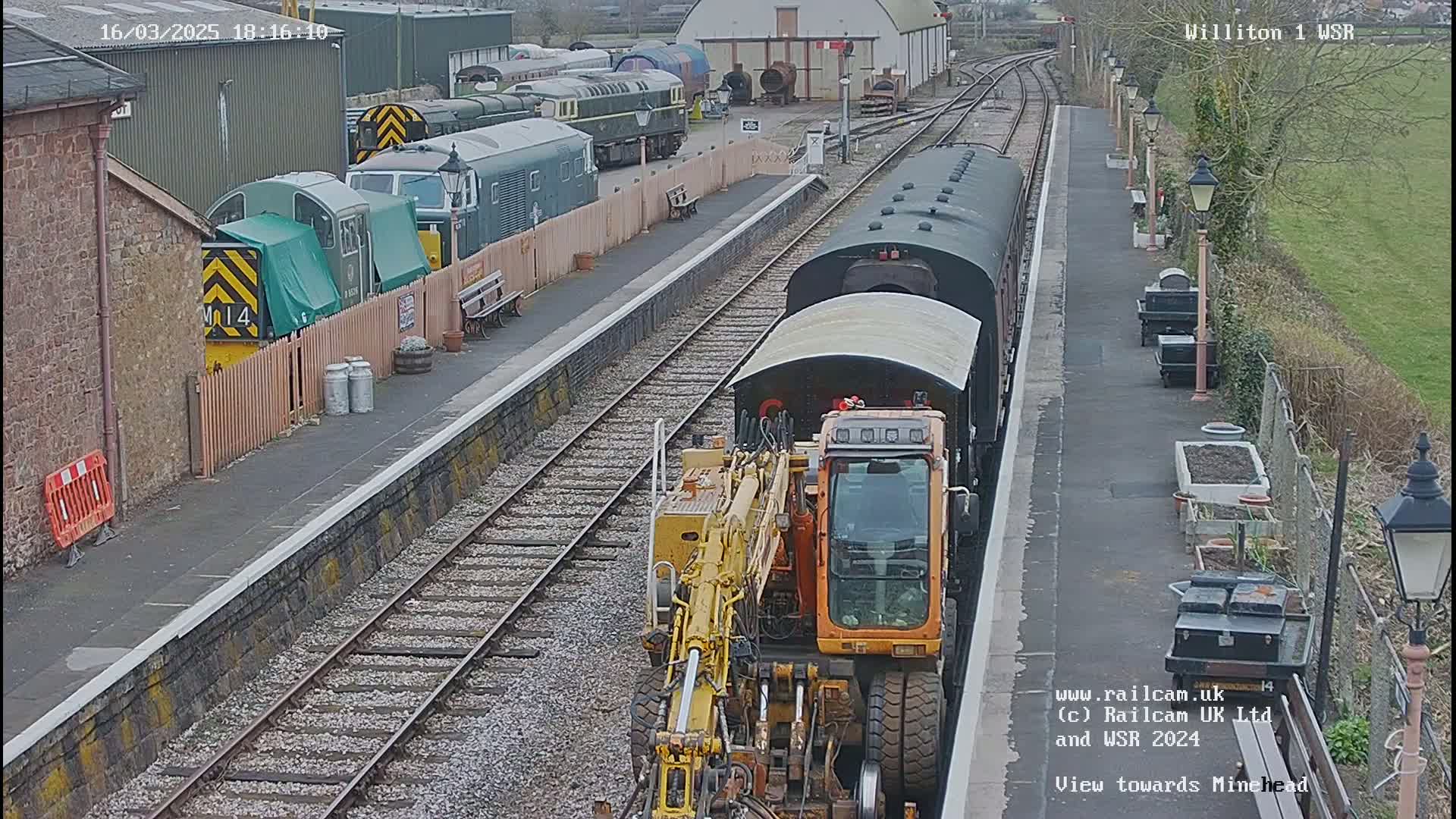 A yellow rail maintenance vehicle is coupled to a dark-colored train at a railway station on an overcast day.