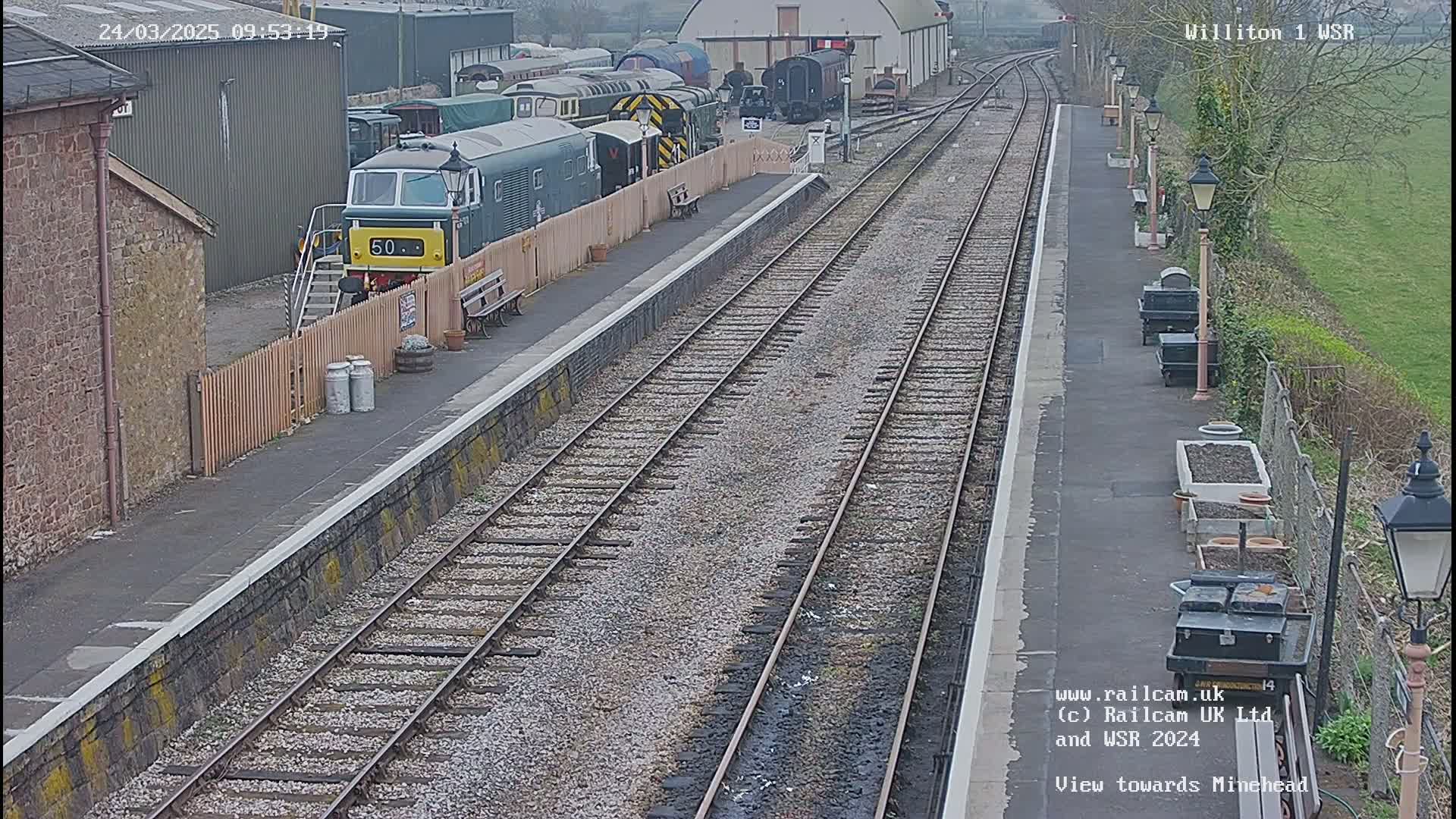 A diesel locomotive is parked at a train station on a partly cloudy day, with multiple train cars visible in the background and railway tracks extending into the distance.