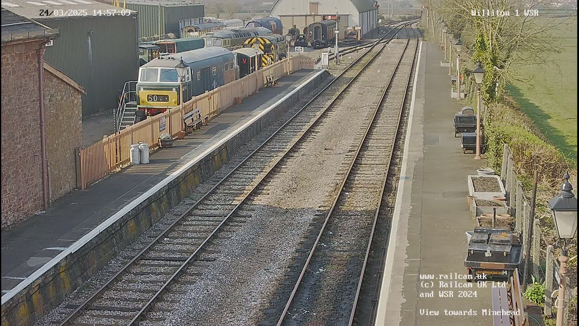A sunny day at a train station shows a diesel locomotive and several railcars parked at the platform, with multiple train tracks extending into the distance.