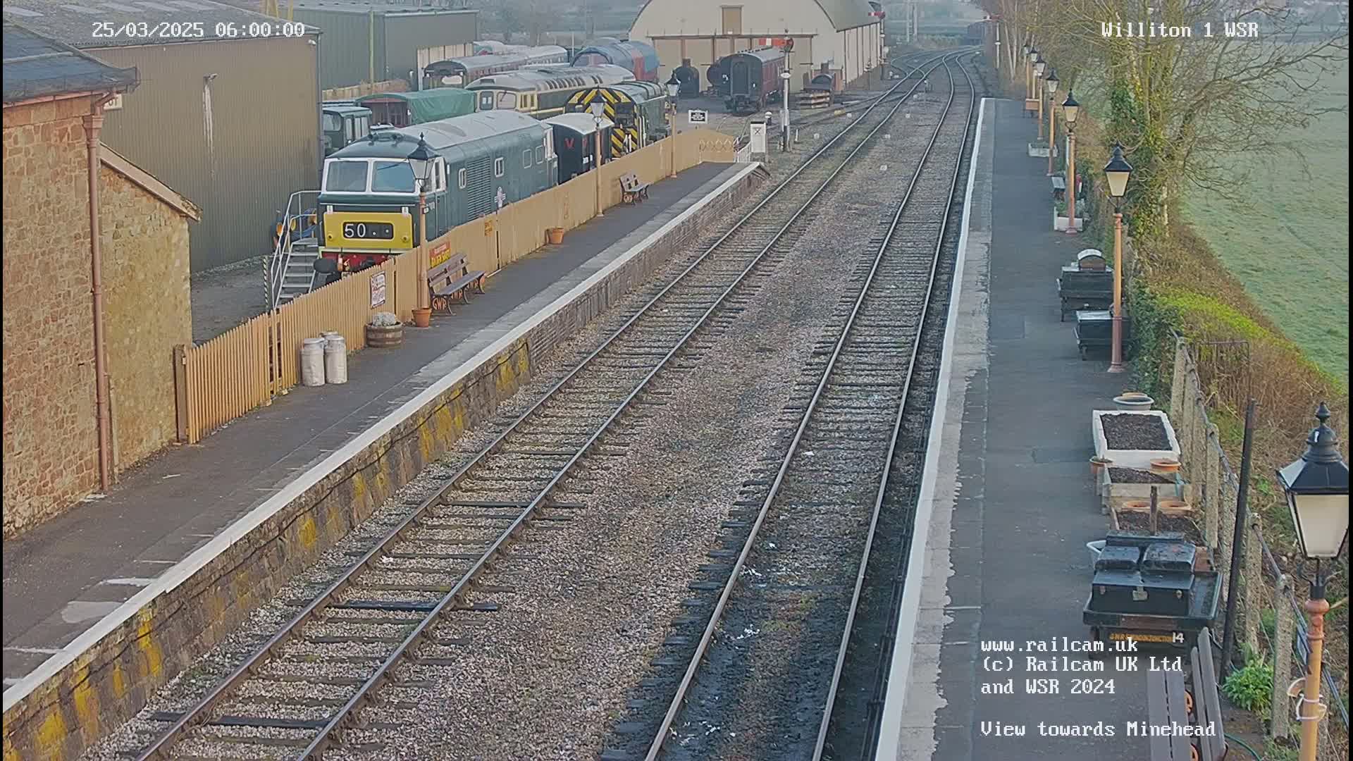 A train sits at a station platform on a slightly overcast morning, with multiple rail lines extending into the distance.