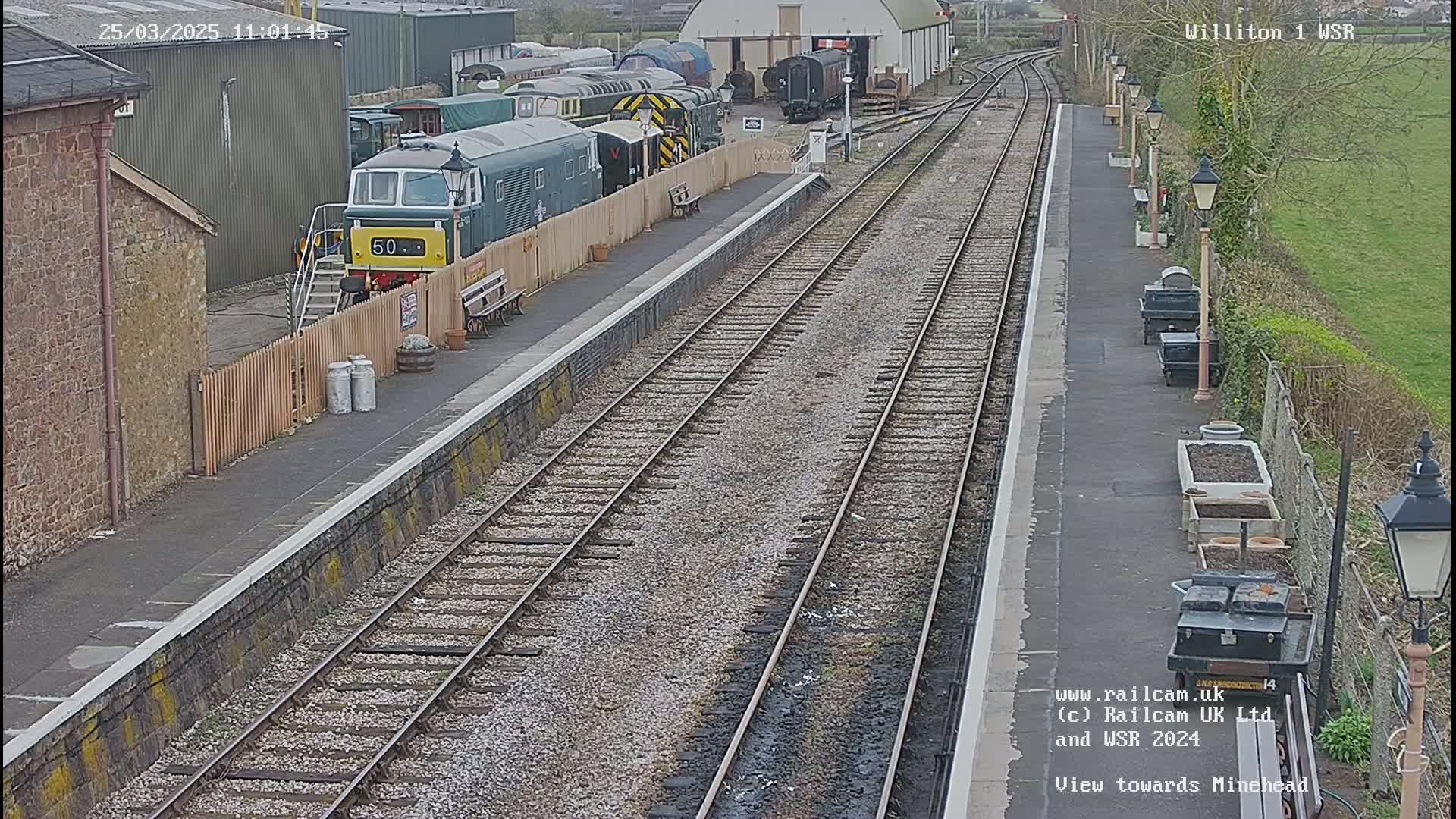 A train station with multiple railway tracks, a diesel locomotive, and several other train cars parked nearby, under an overcast sky.
