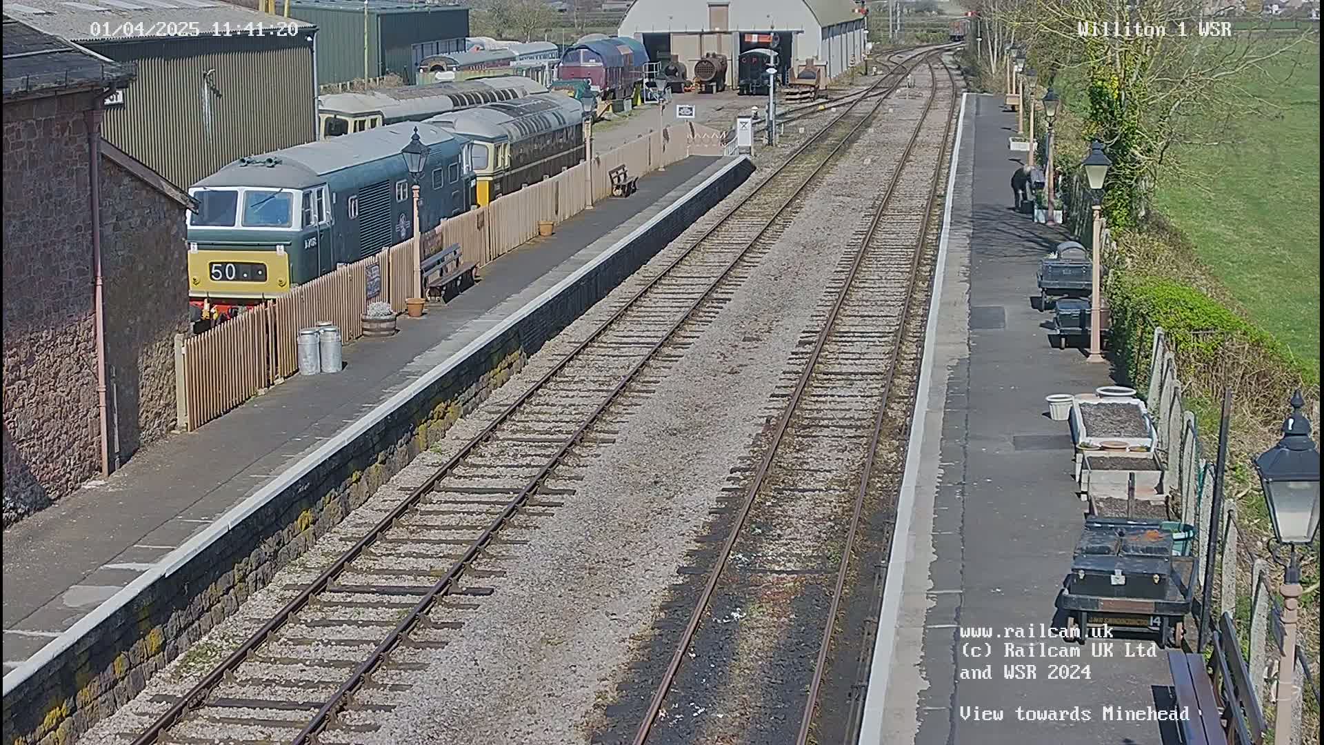 A sunny day at a train station shows multiple train cars parked on a siding next to a platform with two parallel sets of tracks extending into the distance.