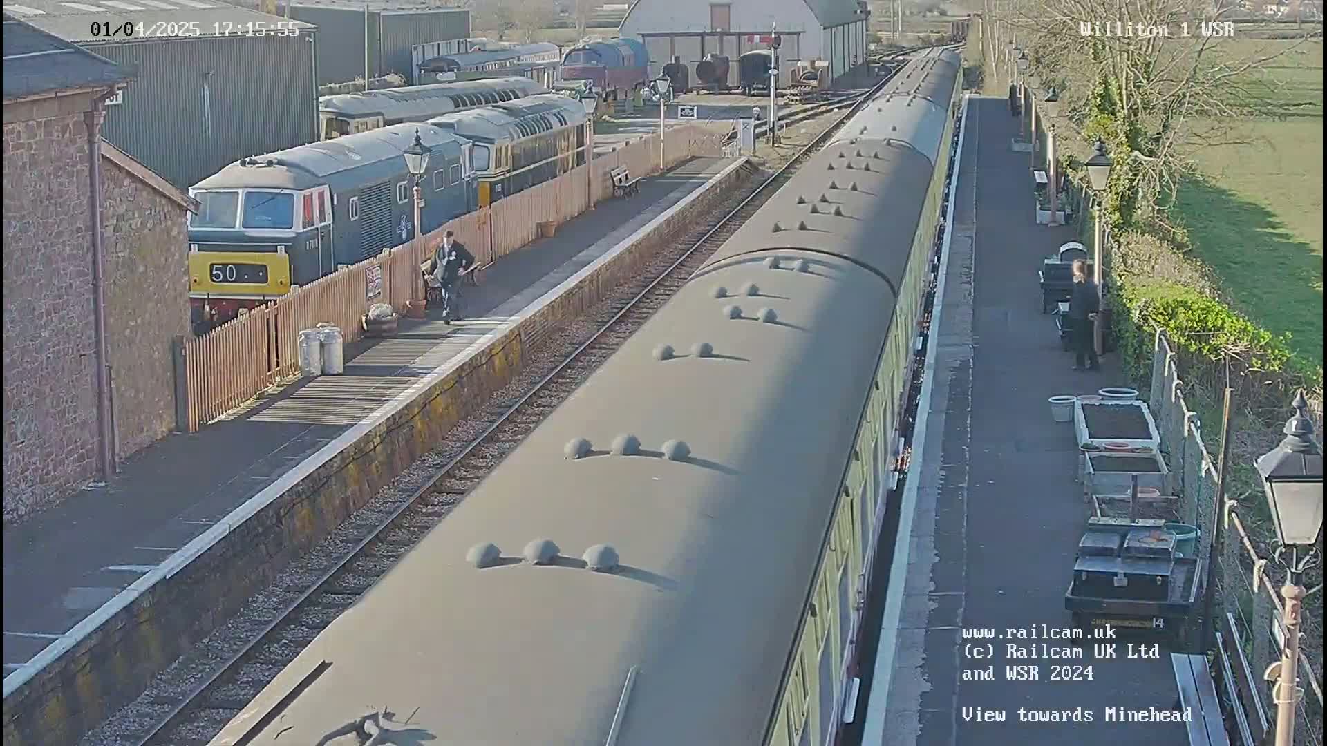 A train pulls into a railway station on a sunny day, with other trains and railway staff visible.