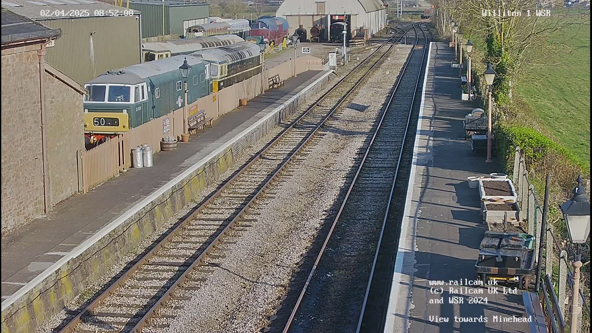 A sunny day at a train station shows multiple train cars and a diesel locomotive parked on a side track next to a platform with two parallel sets of train tracks extending into the distance.