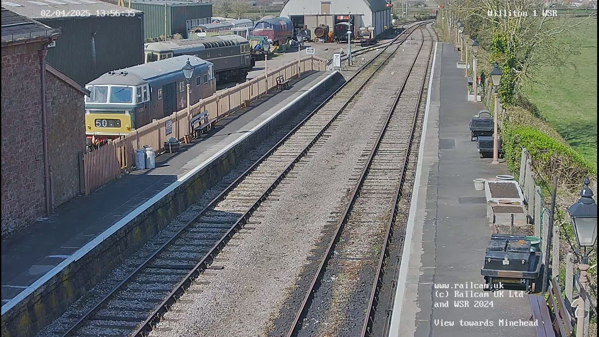 A sunny day at a train station shows a diesel locomotive parked next to a platform with multiple train tracks extending into the distance.