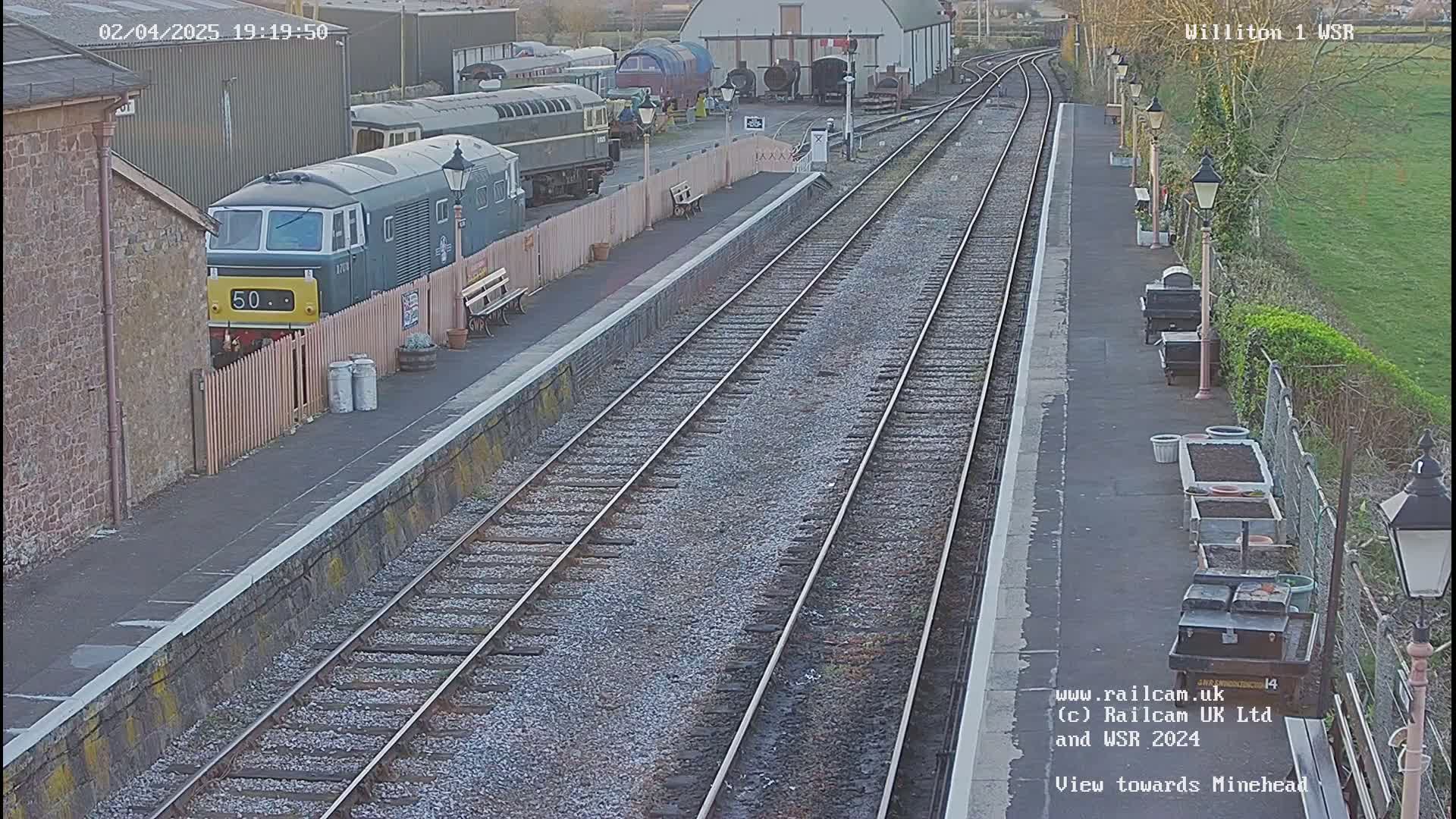 A train sits at a station platform next to other trains and railway buildings, under a twilight sky.
