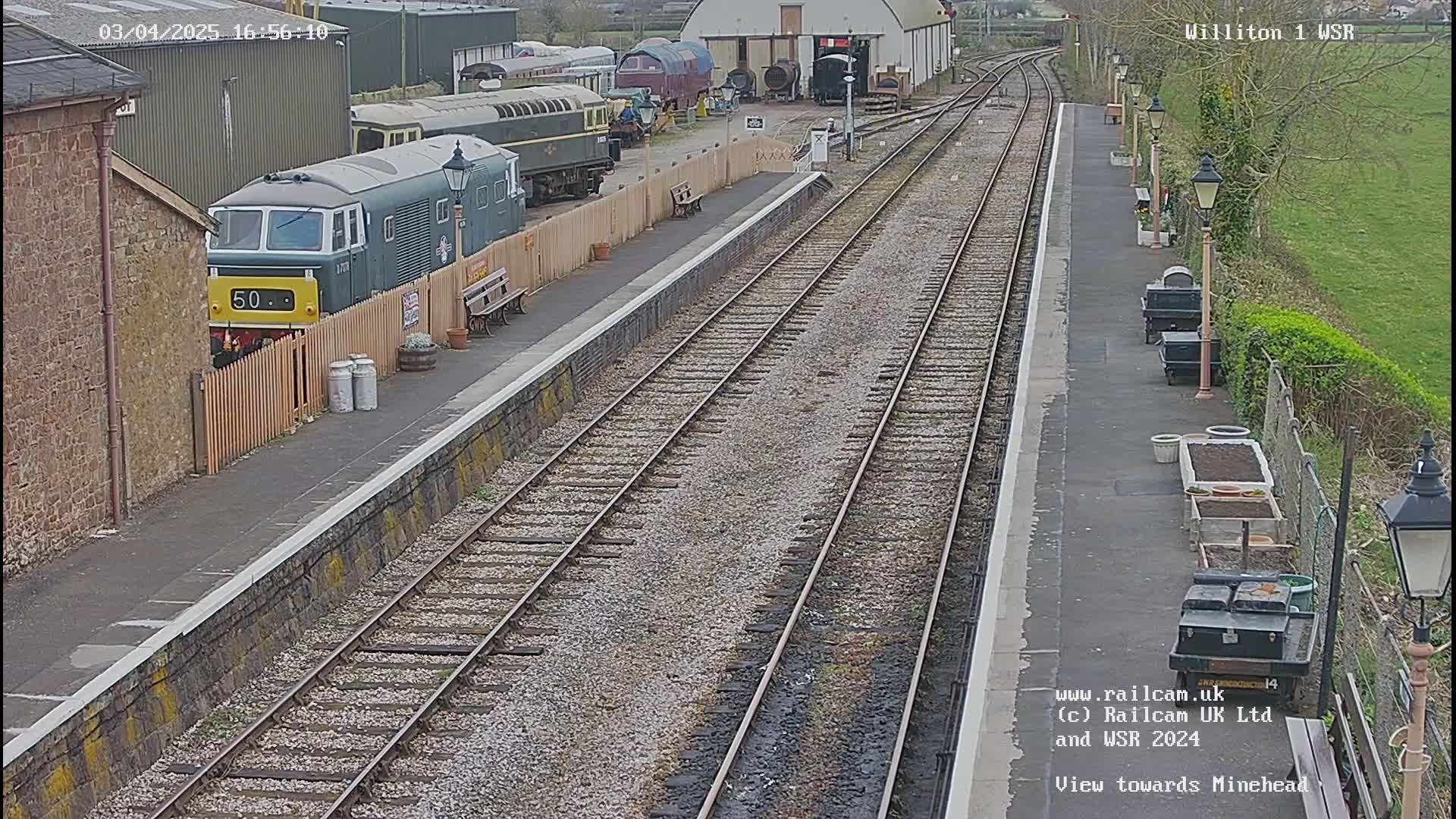 A green diesel locomotive sits at a train station platform next to a railway line on a partly sunny day.