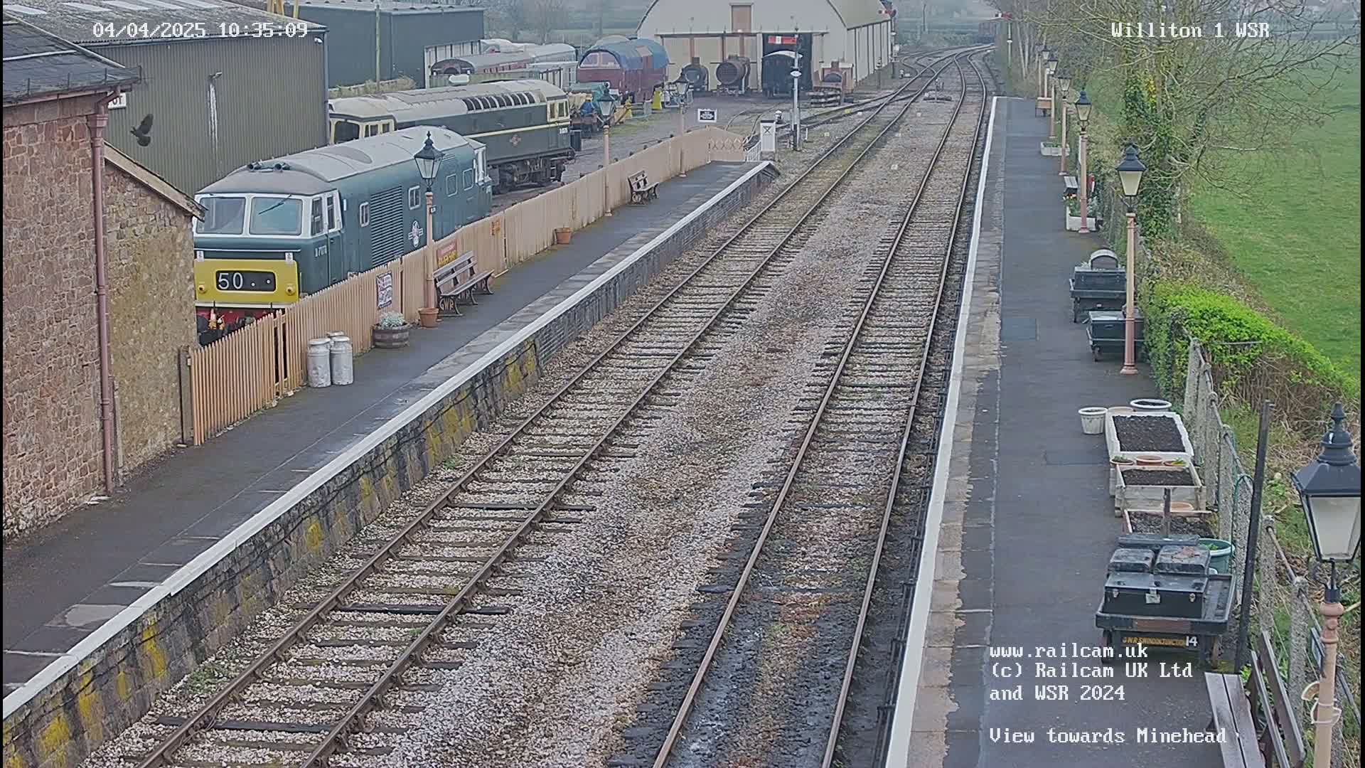 A green diesel locomotive sits at a train station next to several train cars and buildings, on a cloudy day.