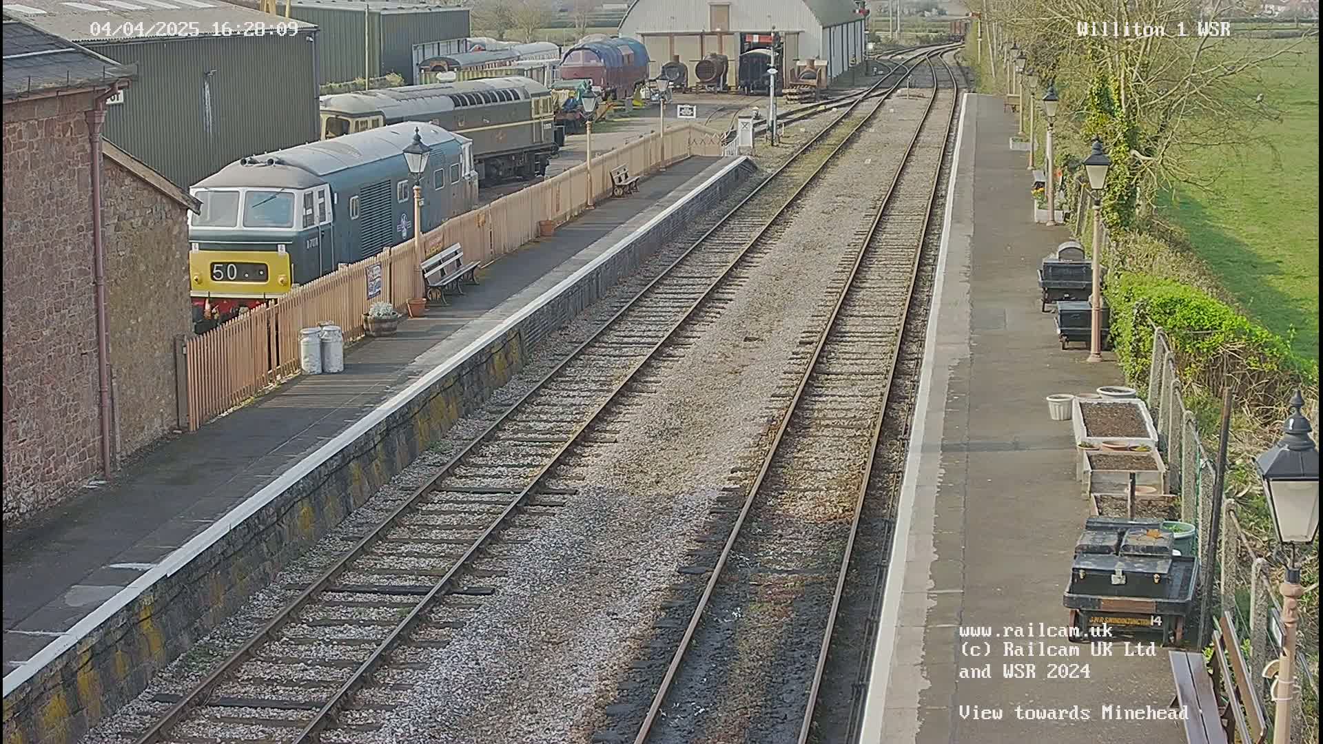 A train sits at a railway station platform on a sunny day, with multiple train tracks extending into the distance.