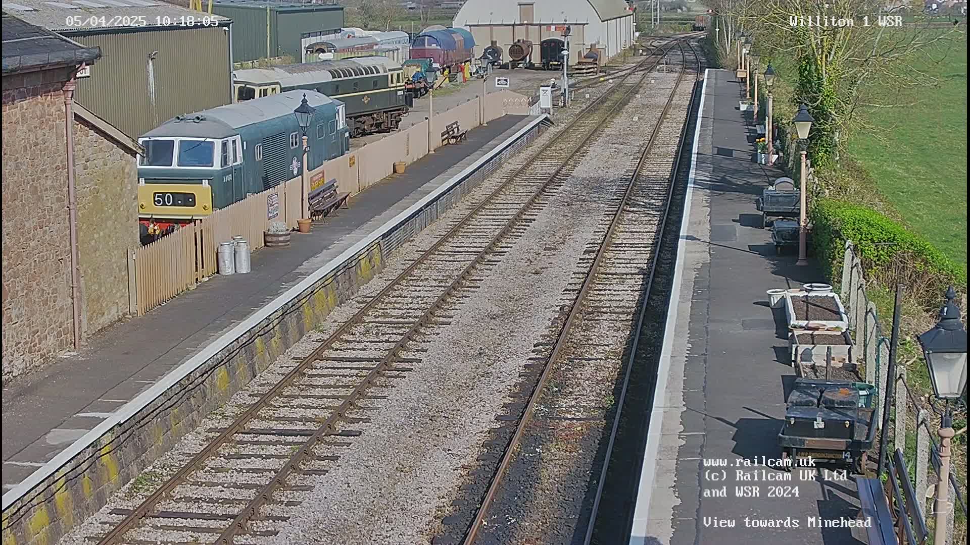 A railway station platform with multiple train tracks, a green diesel locomotive, and several other train cars parked nearby on a sunny day.