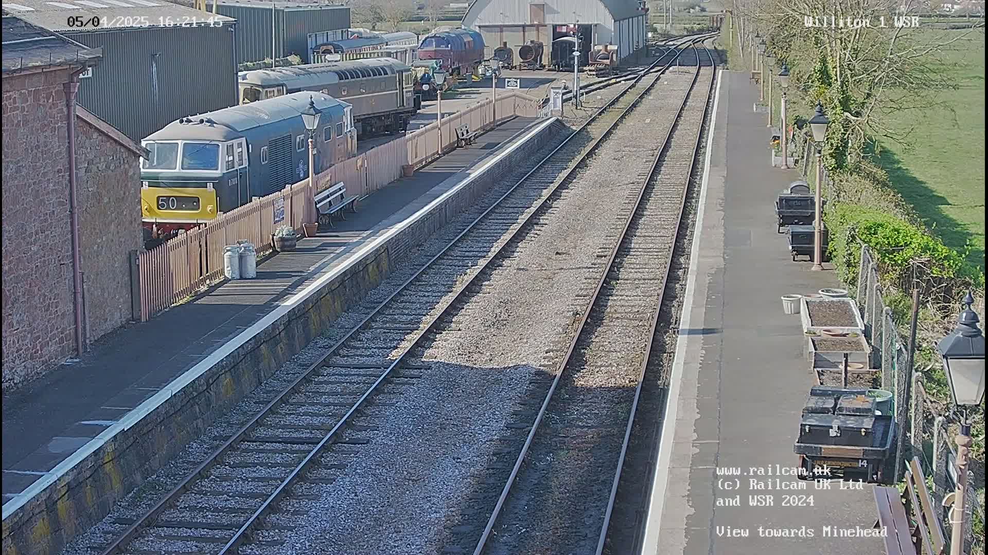 A train sits at a station platform on a sunny day, with multiple train cars visible in the background.