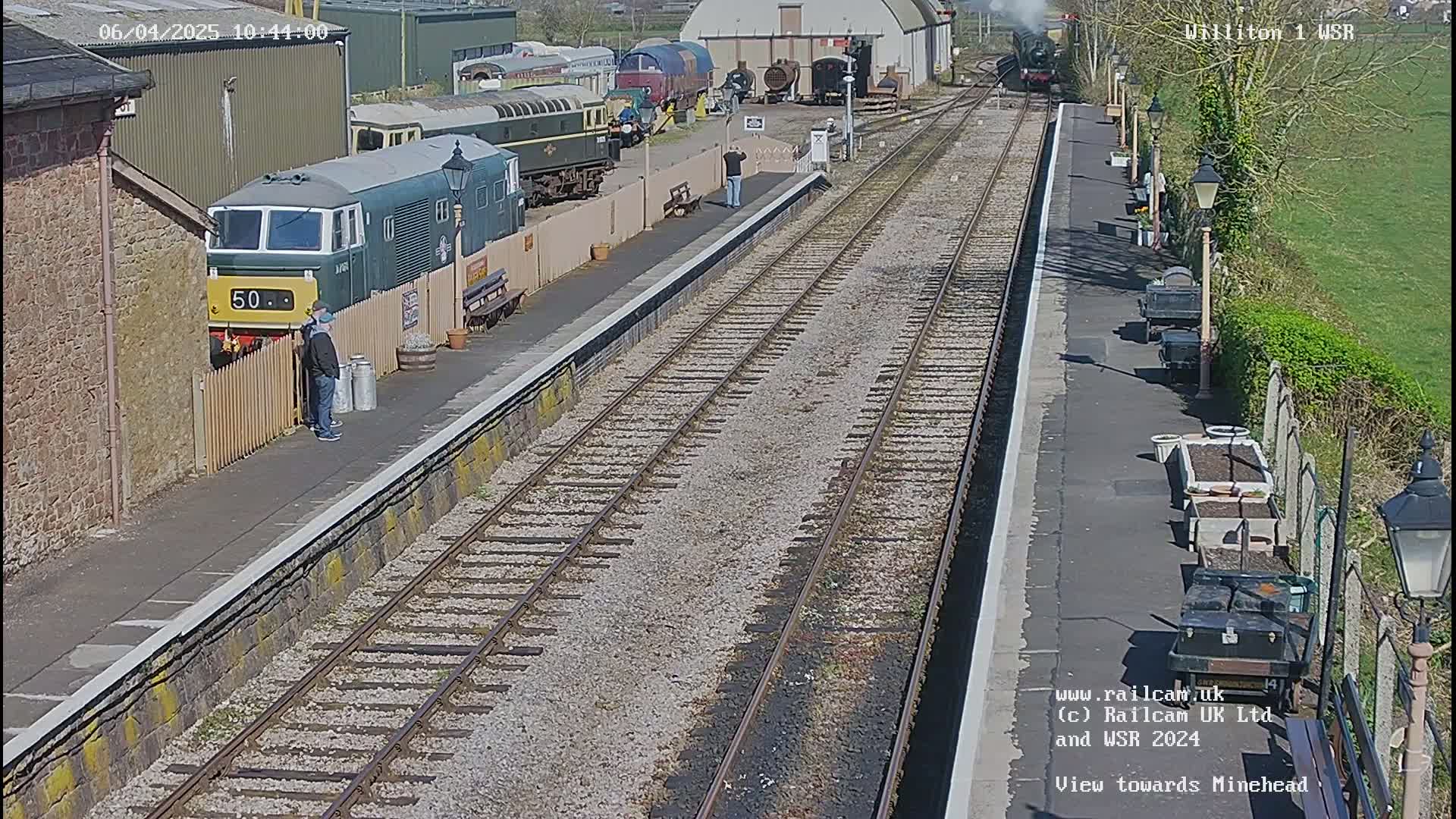 A sunny day at a train station shows a green diesel locomotive parked next to a platform, with multiple railway tracks extending into the distance and other trains visible in the background.