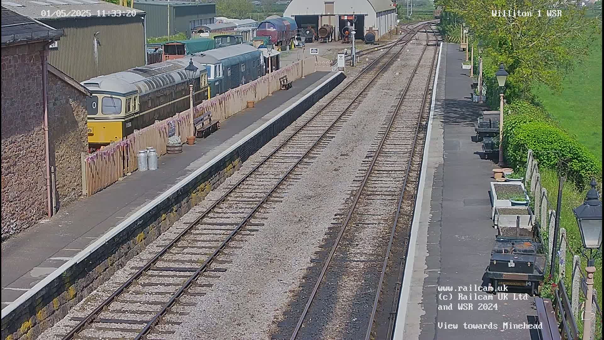 A sunny day at a train station with multiple train cars visible, alongside a platform and tracks extending into the distance.