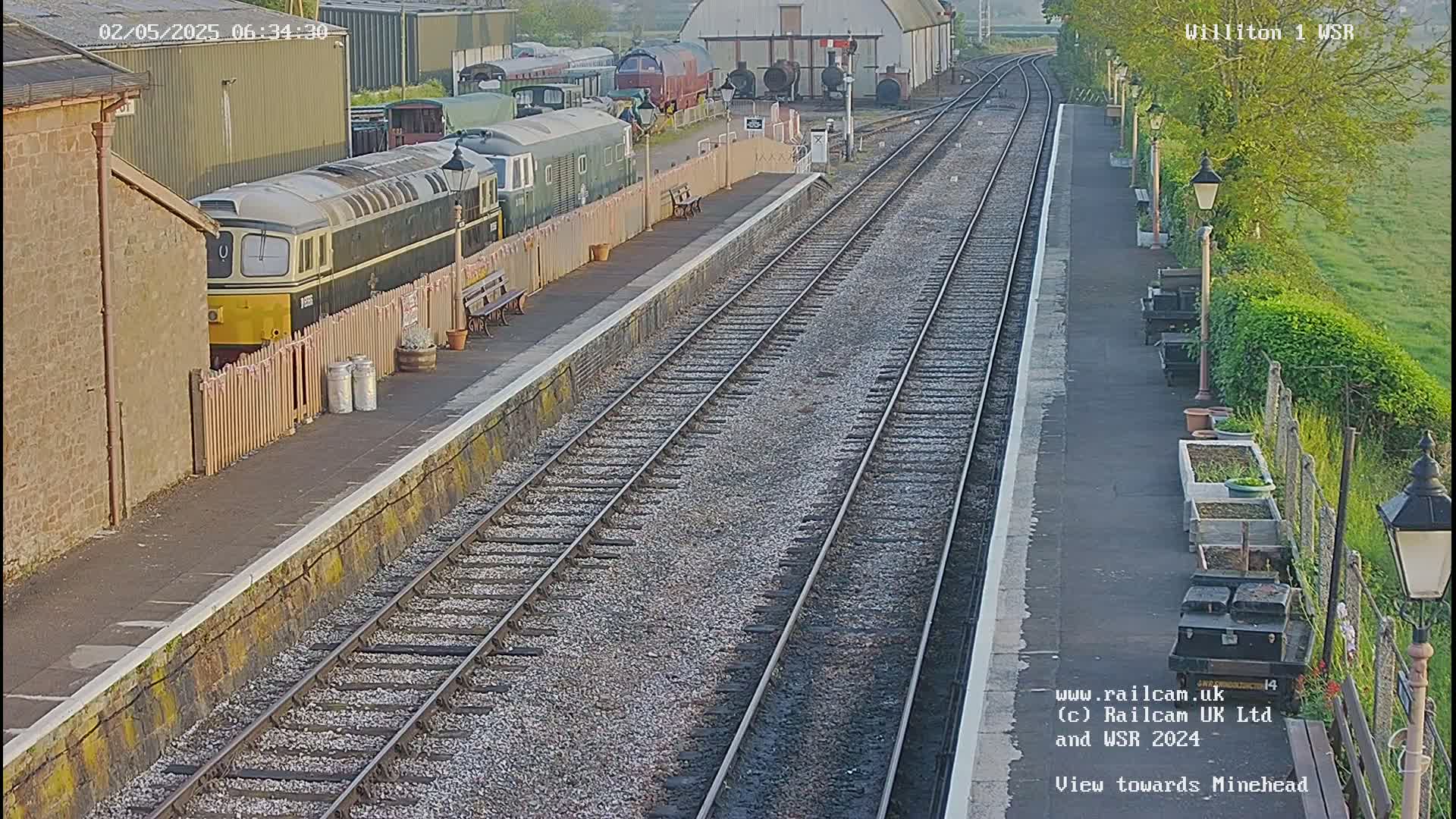 A railway station platform with multiple train tracks, a diesel locomotive, and other railway cars, under a partly cloudy sky.