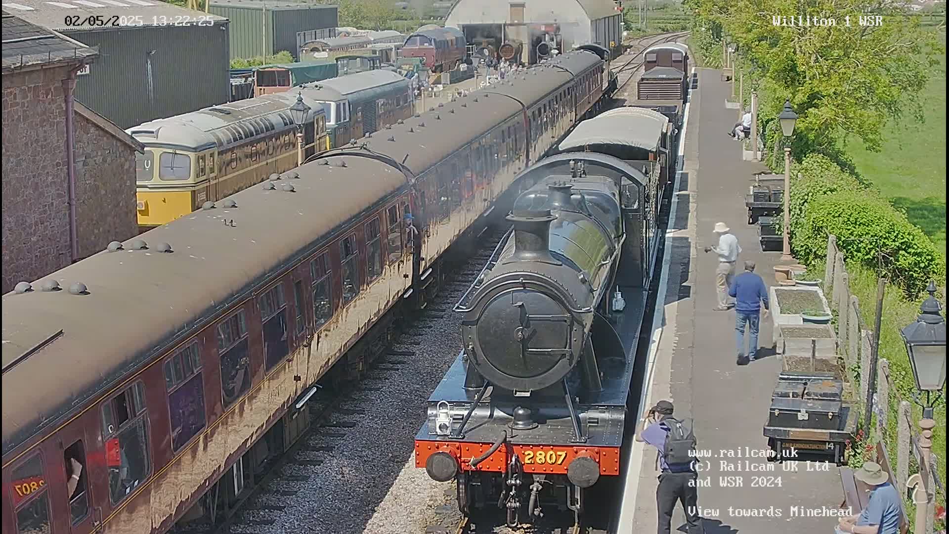 A black steam locomotive and several brown passenger cars are parked at a train station on a sunny day.