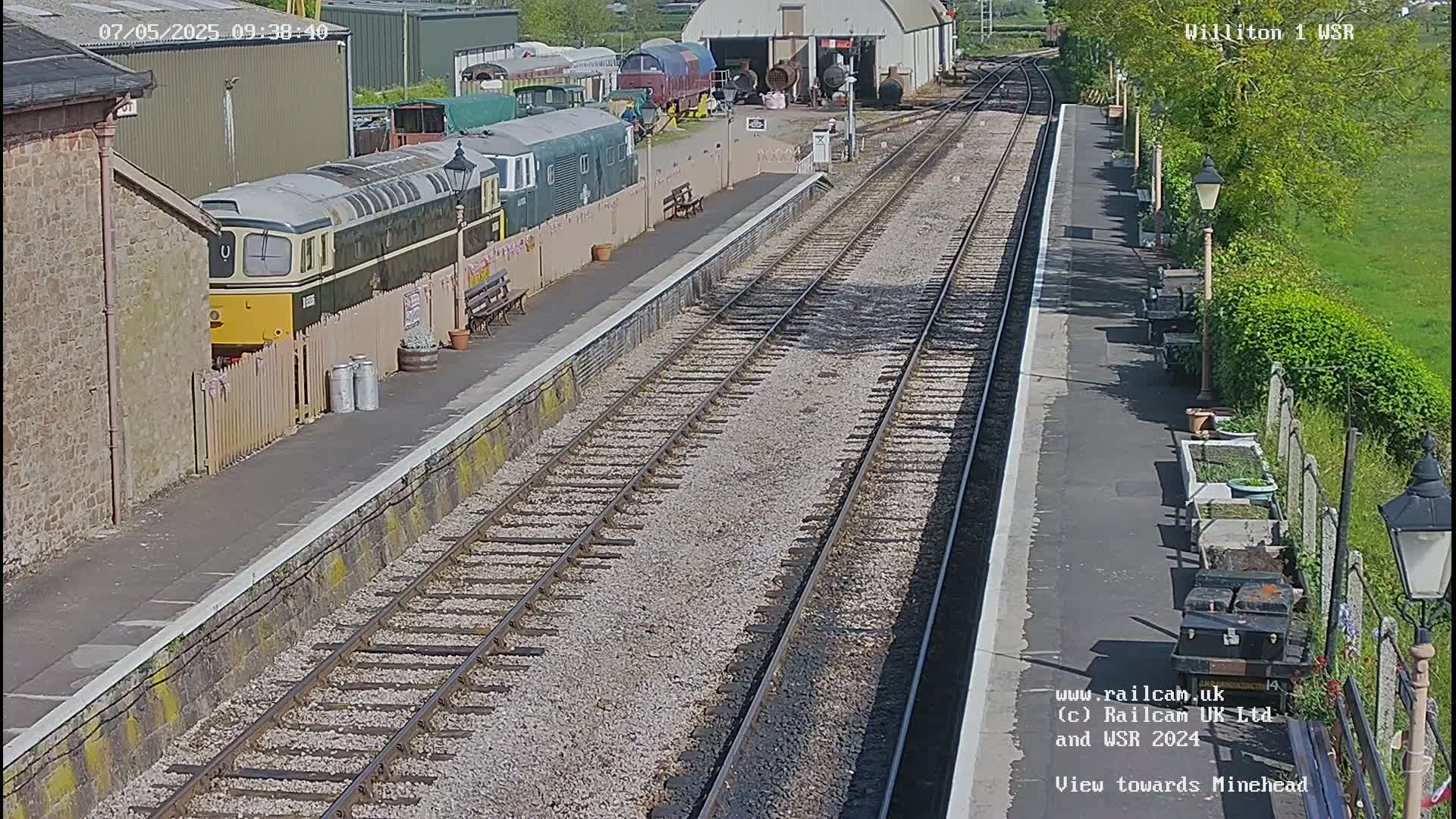 A sunny day at a railway station with multiple train cars and a diesel locomotive parked alongside a platform, featuring parallel railway tracks extending into the distance.