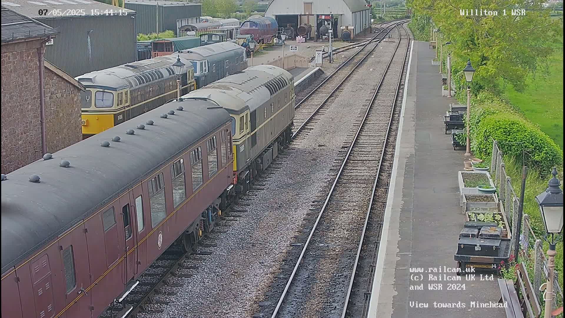 Several diesel locomotives and passenger cars are parked on train tracks at a station on a partly sunny day.