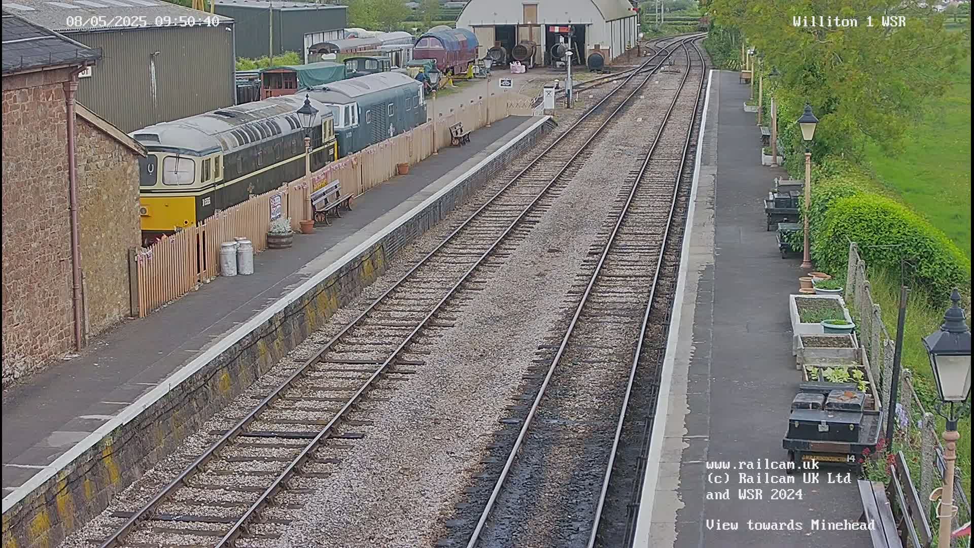 A train sits at a station platform next to railway tracks on a partly sunny day.