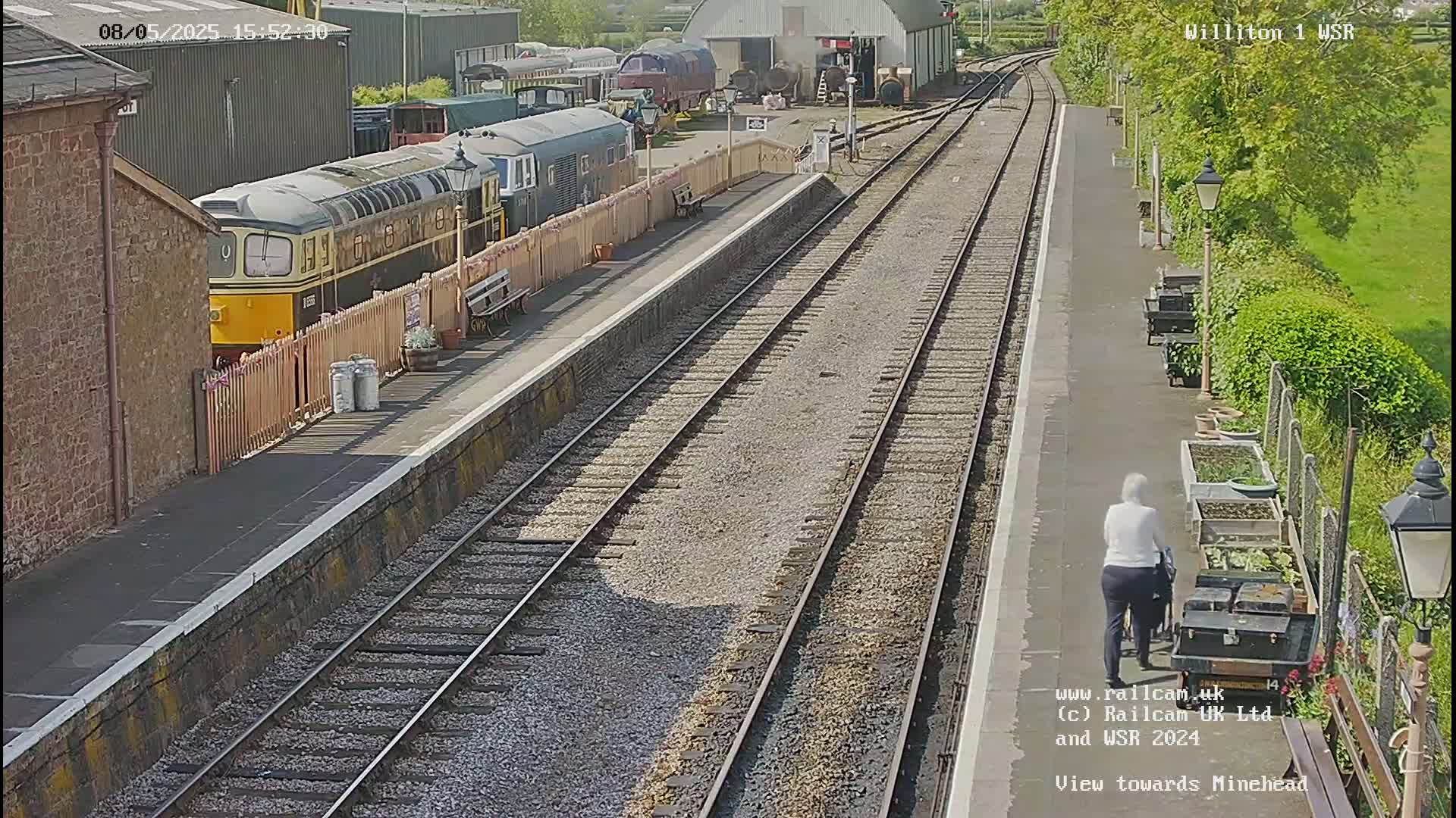 A sunny day at a train station shows a diesel locomotive parked next to a platform with multiple railway tracks extending into the distance, and a person walking on the platform.