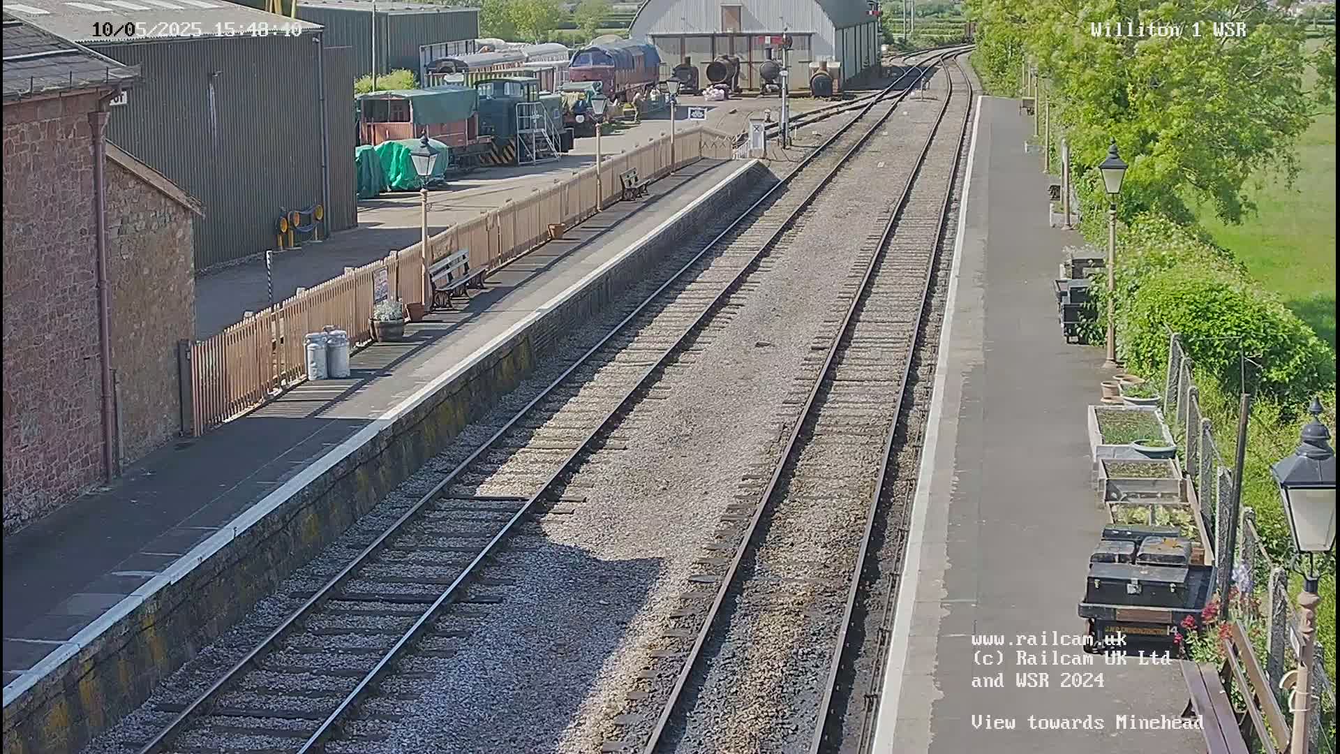 A sunny view of a train station platform with multiple train tracks extending into the distance, alongside a railway yard with several parked trains and railway buildings.