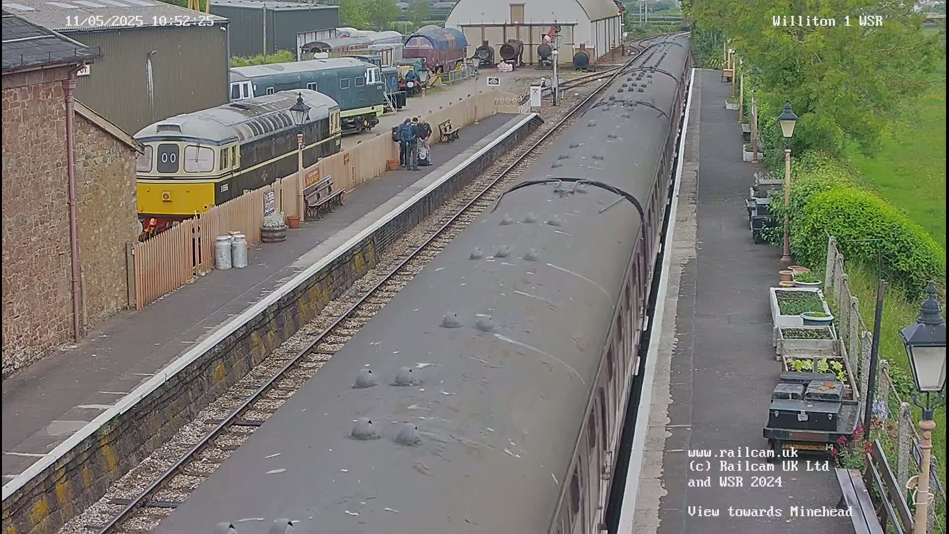 A train is at a station on a partly cloudy day, with several people standing nearby and other railway cars visible in the background.